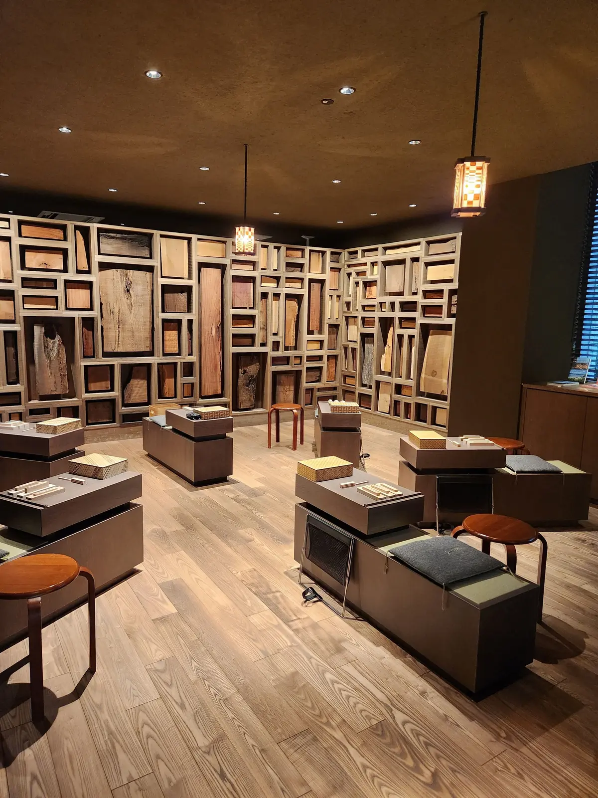 木目調の床と壁面収納が特徴的な展示空間。壁面には木材のサンプルが並び、中央には立方体の展示台が配置されている。照明は天井埋め込み型と吊り下げ型で、温かみのある光が全体を照らしている。