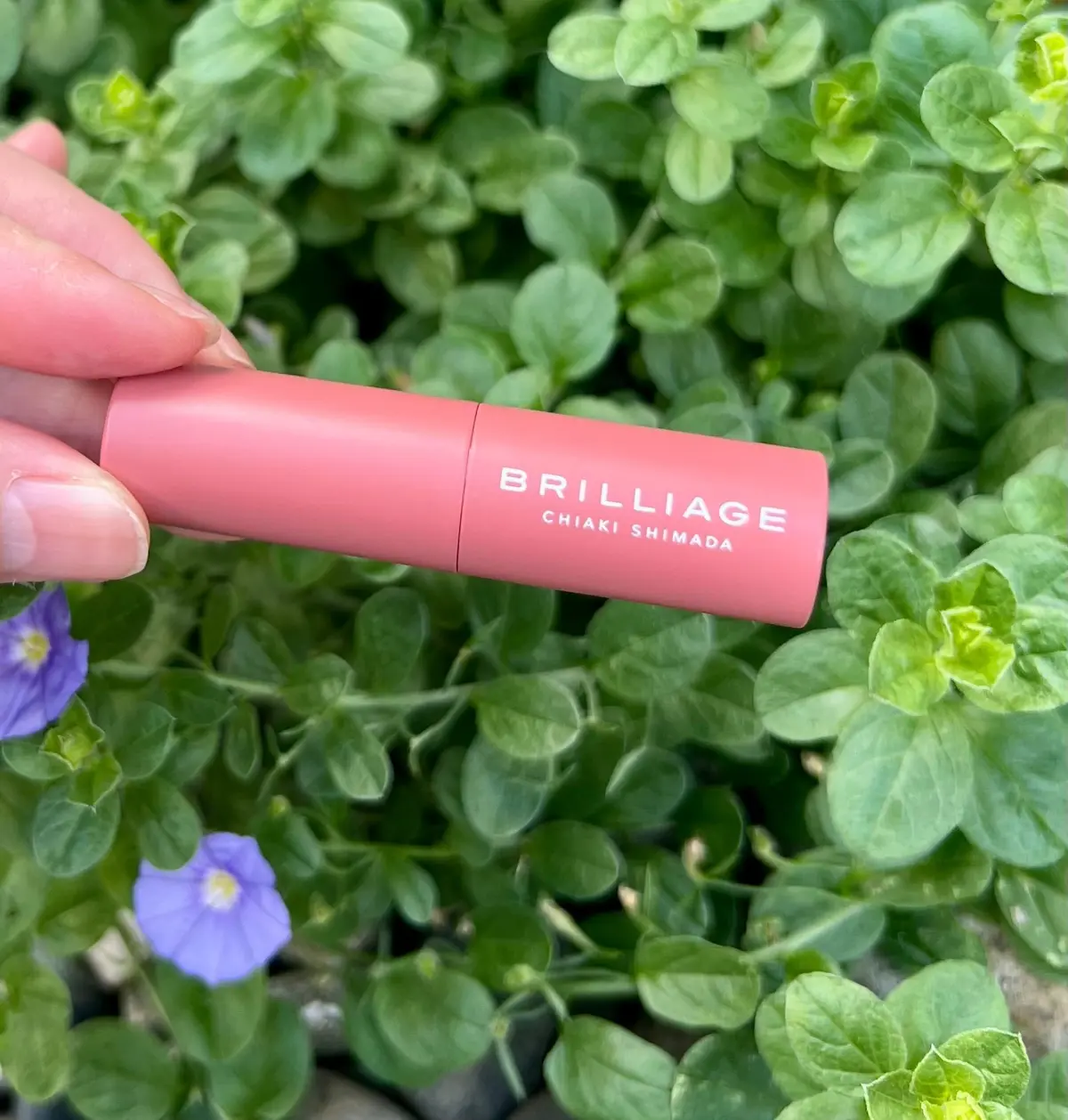 ピンクの化粧品チューブ「BRILLIAGE CHIAKI SHIMADA」を手で持ち、緑の低木と青い花の背景に置かれています。