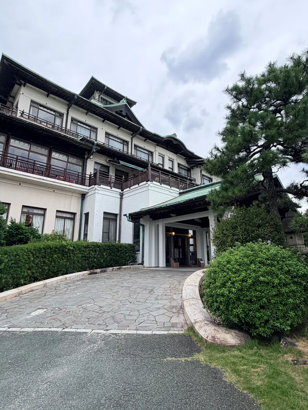 白とクリーム色の建物と、緑豊かな植栽が特徴的な風景。入り口は屋根付きで、石畳の道が続いている。空は曇り。