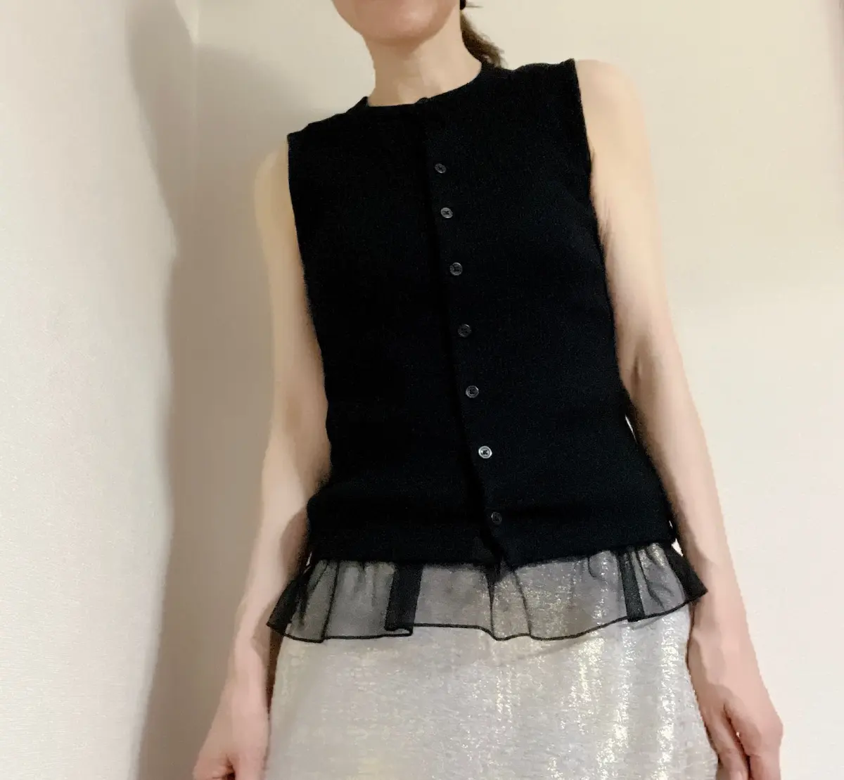 黒いノースリーブのトップスとシルバーのスカートを着用した女性。トップスは丸襟で、前側にボタンが並んでいる。裾はフリルになっている。