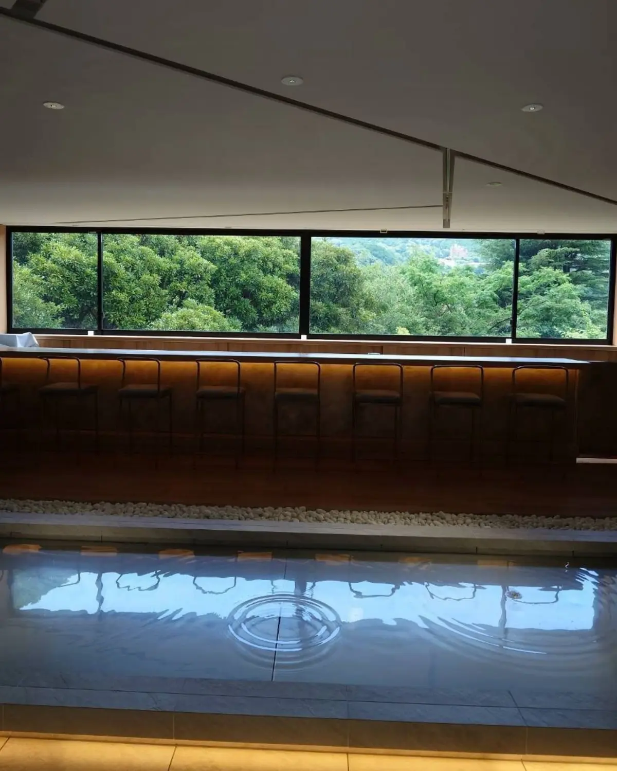 三輪湯河原のバー。木目調のカウンター席と、その奥の大きな窓から見える緑豊かな風景。カウンターの下には白い小石が敷き詰められ、足元には水面に円形の波紋が映る水盤。黒いスツールが並ぶ、明るい空間。