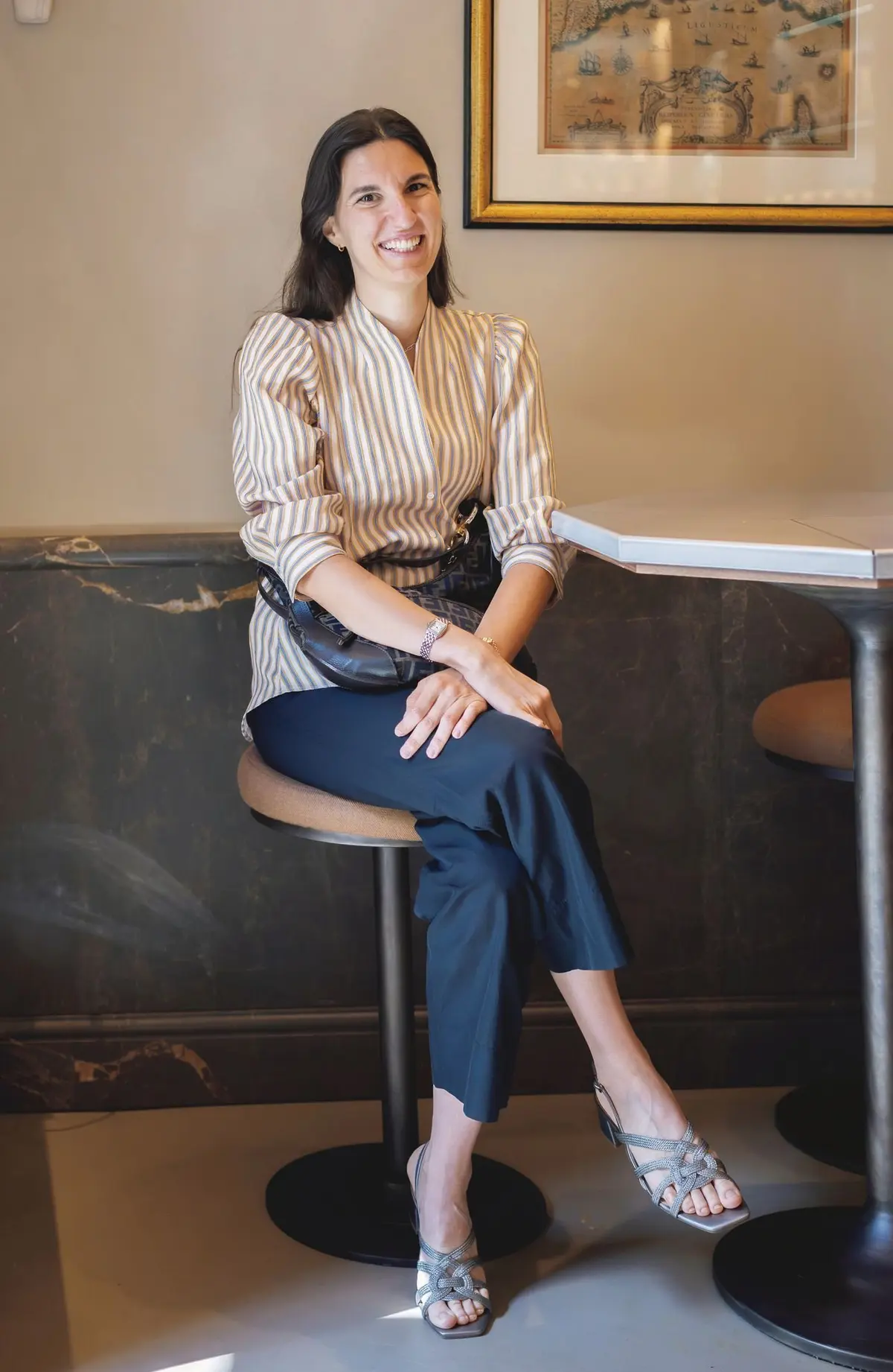 明るい笑顔の女性が、丸いスツールに座ってこちらを見ている。ストライプのシャツに紺色のパンツ、シルバーのサンダルを着用。背景には、模様の入った壁と絵画が飾られている。