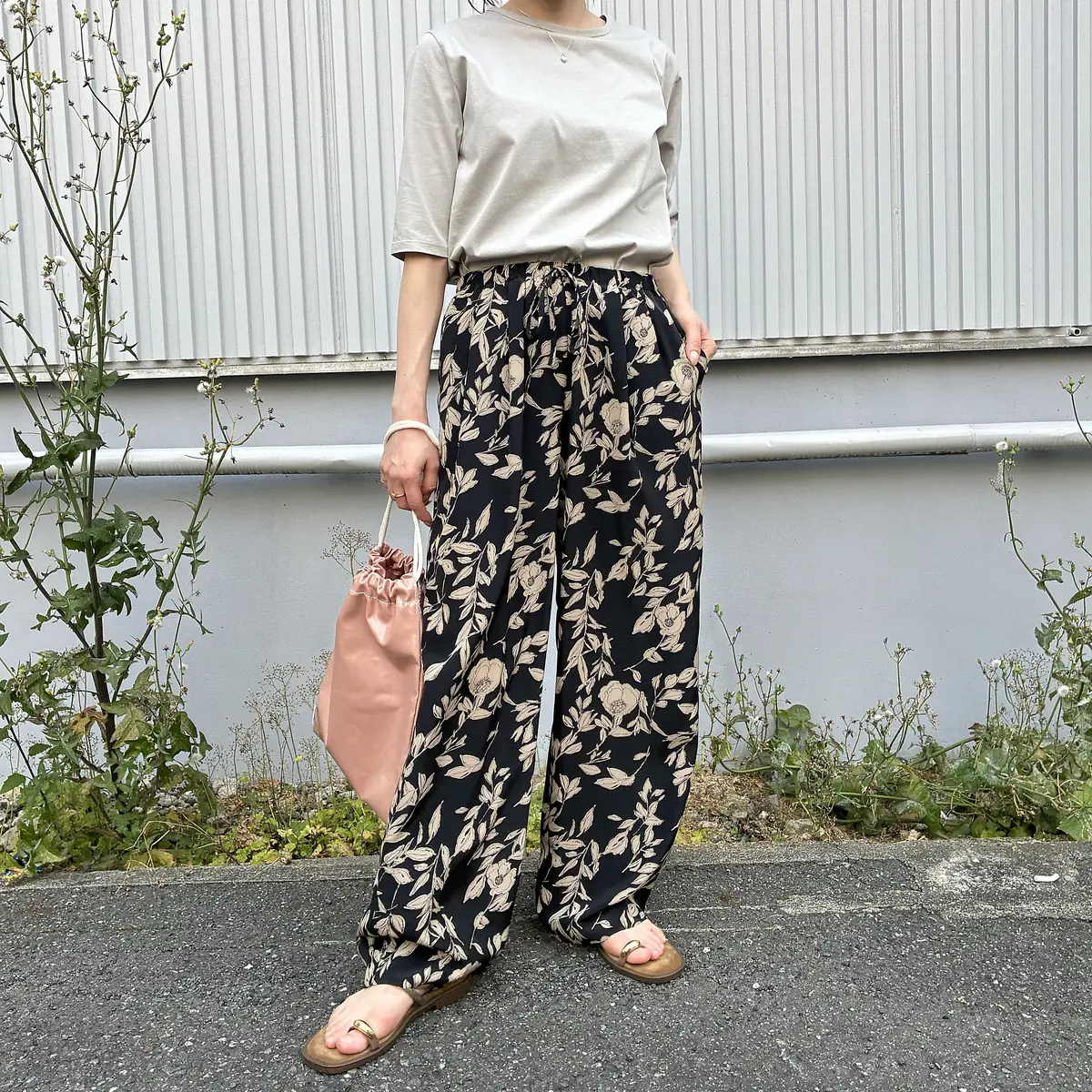 ベージュの半袖トップスと黒の花柄ワイドパンツを着た女性が、ピンクのトートバッグを左手に持ち、コンクリート地面に立っている。背景には緑の植物と白い壁がある。足元は茶色のサンダル。
