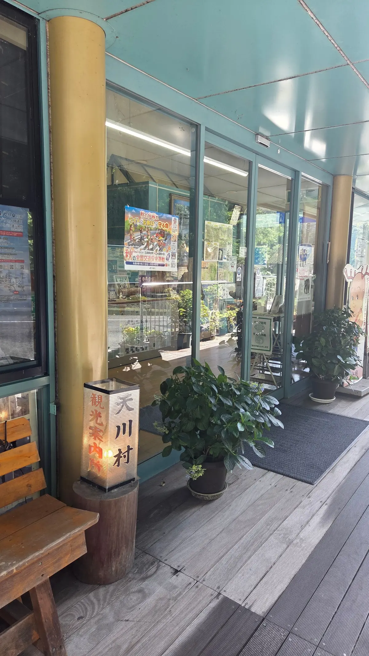 木造のデッキに面した、淡い水色の店舗の入り口。右には観葉植物が置かれ、左には木製の椅子と、縦長の看板が立っている。看板には日本語で「天川村 観光案内 観光案内所」と書かれている。