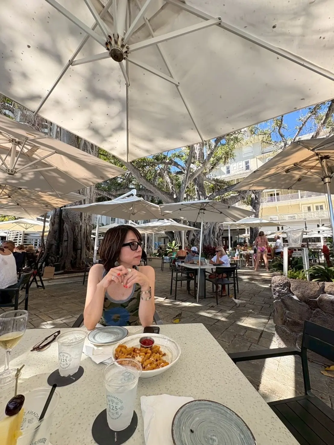 屋外レストランのテラス席で、女性が食事を食べている。テーブルにはワイン、飲み物、ポテトの盛り合わせが置かれ、周囲には傘付きのテーブルや木々が広がる。