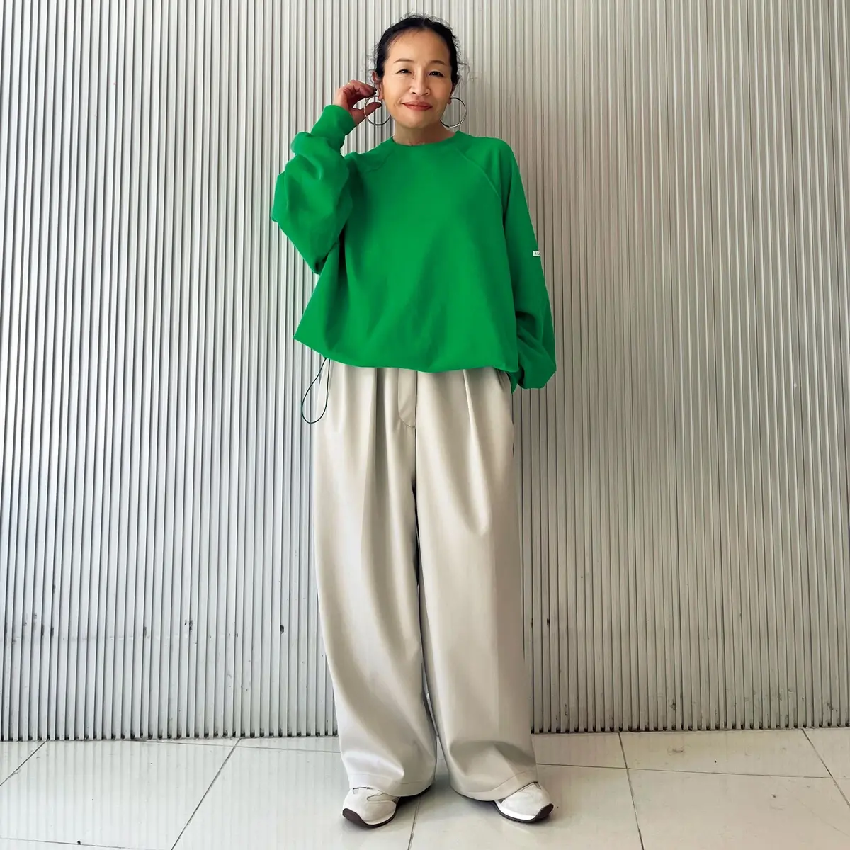 緑色のオーバーサイズスウェットとベージュのプリーツパンツを着用した女性。右手を頭に添え、正面を向いている。背景は縦のラインが入った白い壁。
