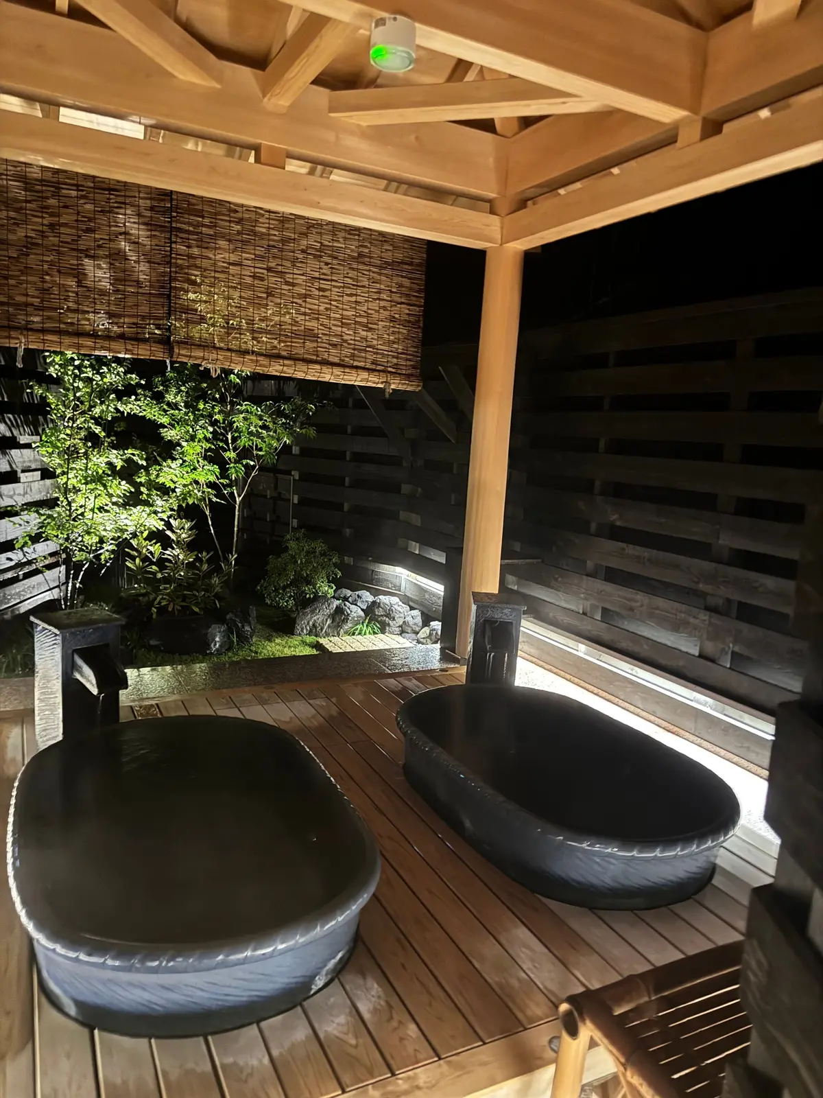 木造の屋根付き露天風呂。2つの黒い湯船が並び、手すりが設置されている。背景には竹垣と植栽が見え、ライトアップされている。
