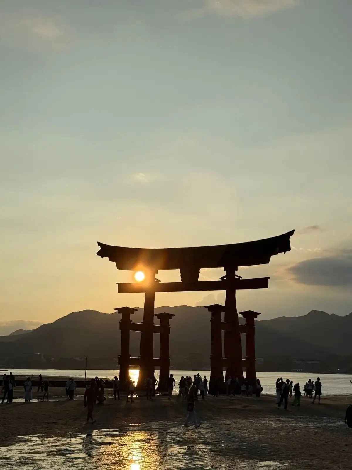 瀬戸内海の夕日に照らされた、厳島神社の鳥居。水平線に沈む太陽が鳥居の輪郭を照らし出し、水面には反射が映る。多くの人がその景色を見ている。