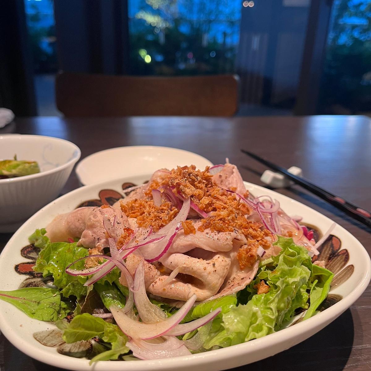 白磁の皿に盛り付けられた豚肉のサラダ。レタスを敷き詰め、薄切りにした豚肉と紫玉ねぎを散らし、砕いたナッツがトッピングされている。