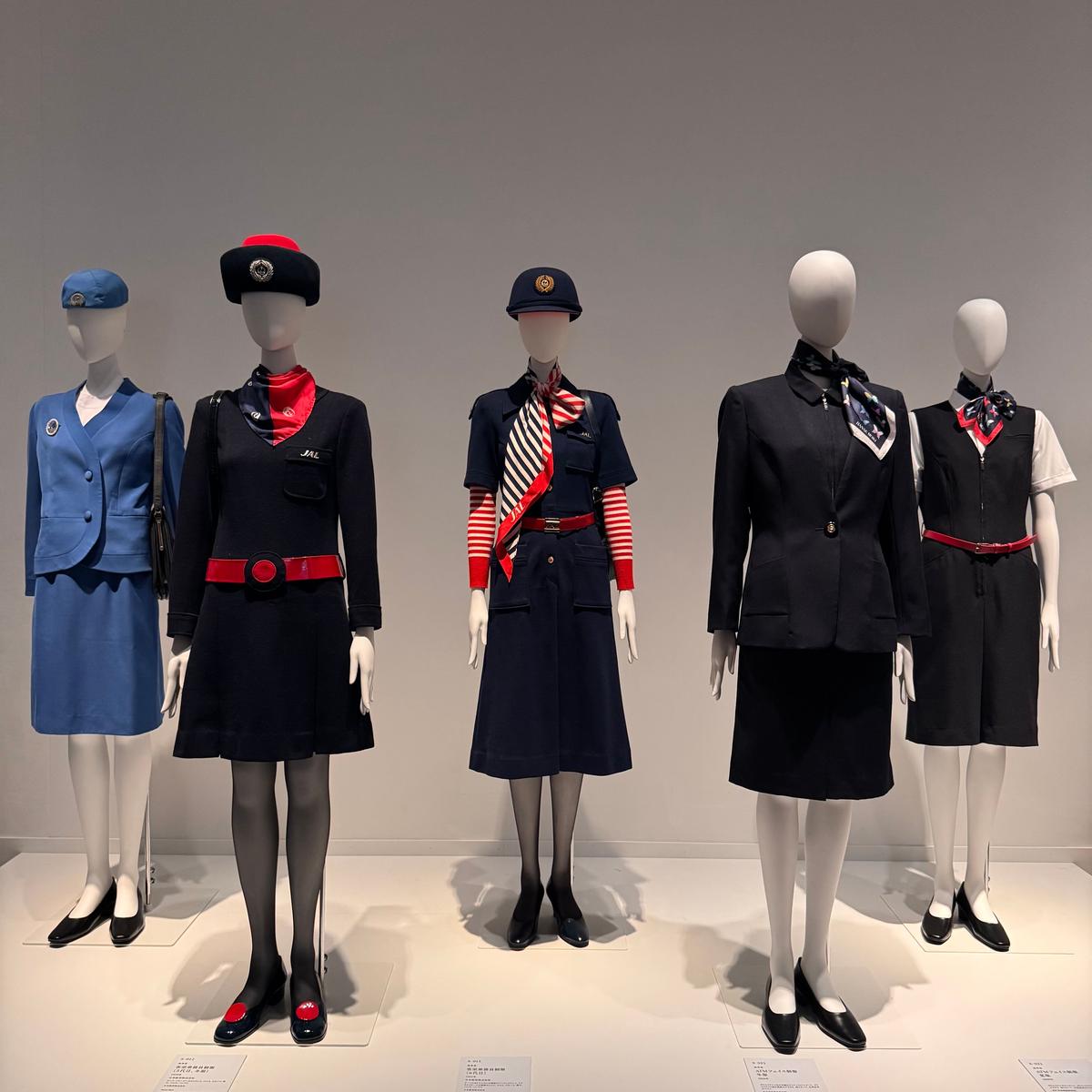 JALの歴代客室乗務員制服展示。6体のマネキンが年代ごとの制服を着用。紺、黒を基調としたスーツやスカート、帽子、スカーフなど。各マネキン前には説明書きがある。