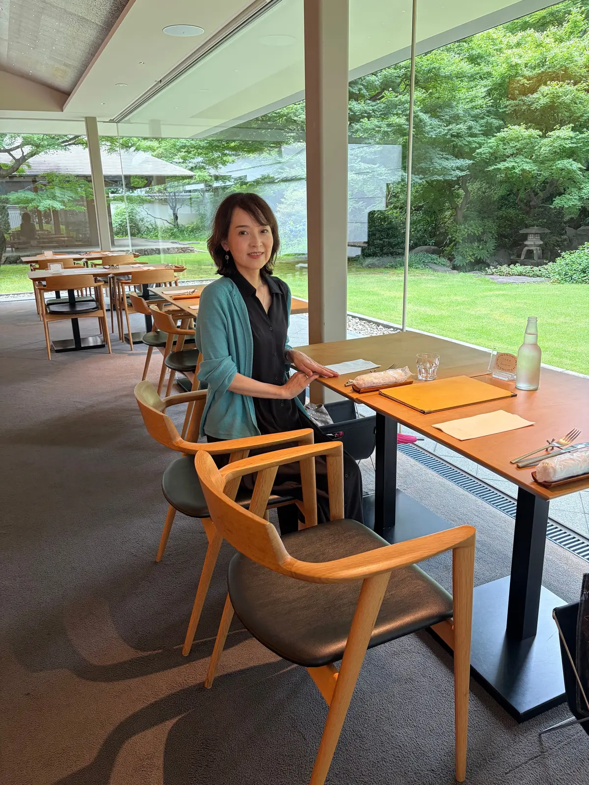 女性が木製のテーブルに座り、明るいカフェのインテリアと緑豊かな庭園を背景に笑顔でポーズしている