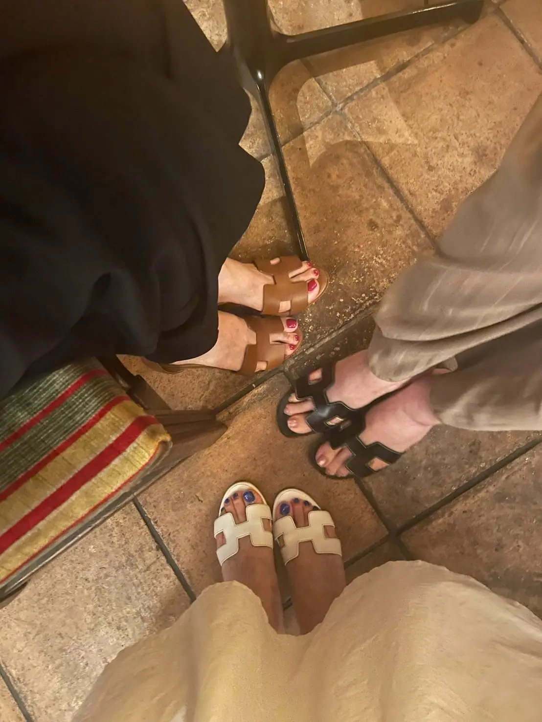 テーブル席で、3人の女性が異なるデザインのサンダルを履いている様子。左から黒いパンツ、ベージュのパンツ、黄色いスカートの順に並び、それぞれ茶色のレザーサンダル、黒いシンプルなサンダル、エルメスのサンダルを履いている。
