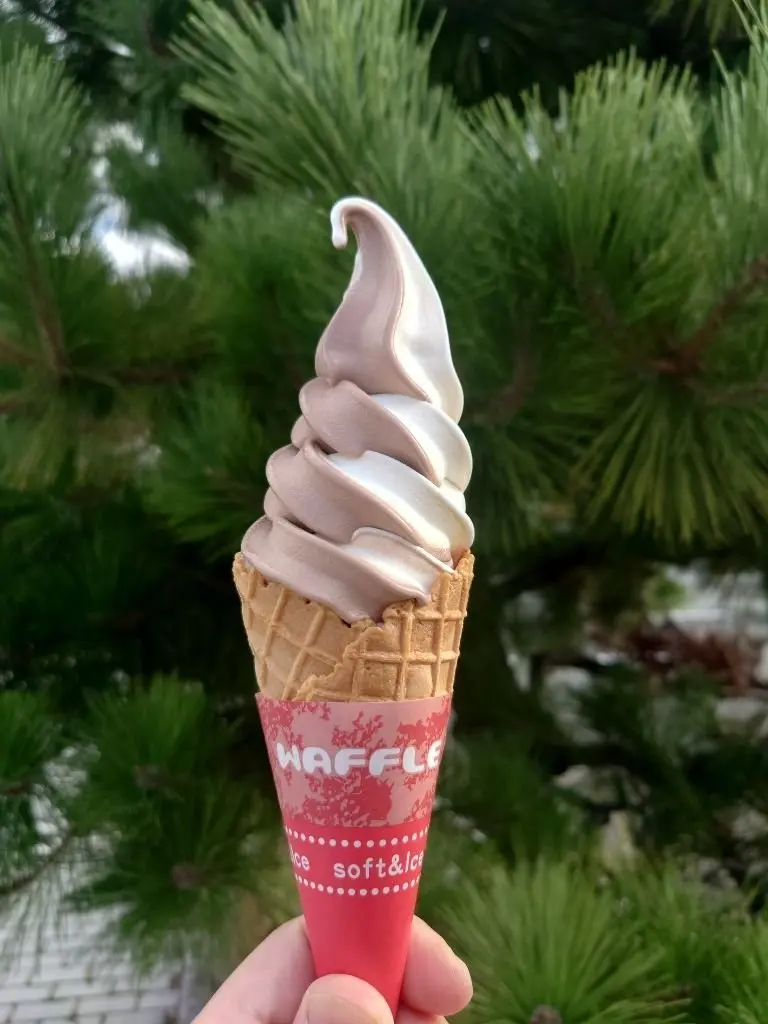 手で保持されたソフトクリームの画像。ワッフルコーンに、白と薄いピンク色の2色のフレーバーがねじり状に盛られている。背景には緑の木々が茂っている。