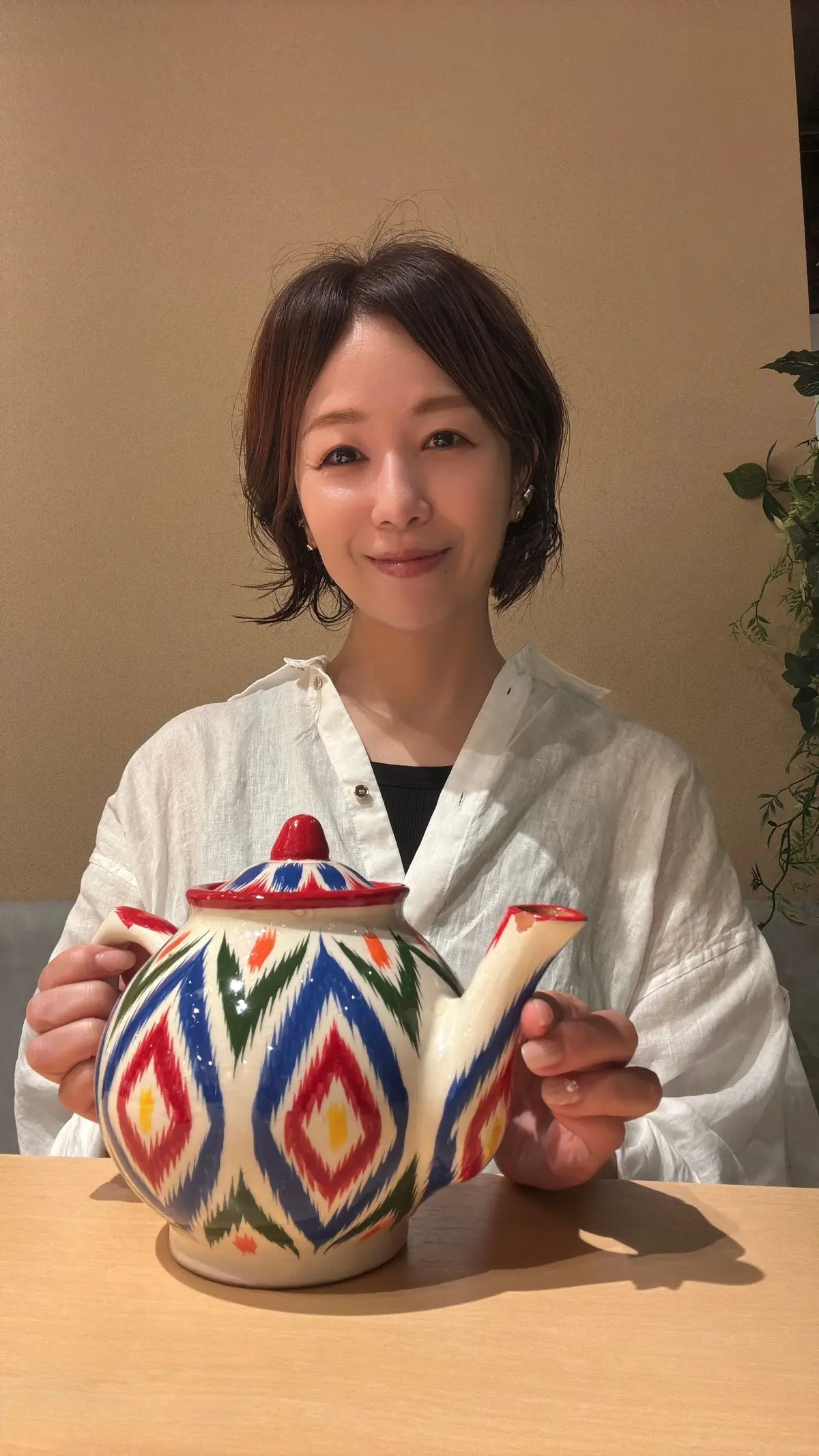 女性が赤と青の幾何学模様が描かれた陶器のティーポットを両手で持ち、笑顔でカメラに向かって見つめている。背景には緑の植物とベージュの壁がある。