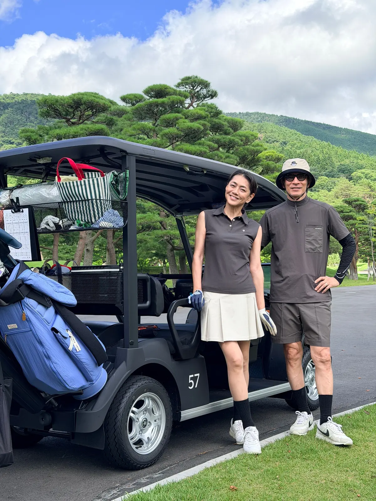 ゴルフカートの前に立つ前田典子さんと日比野怜さん。前田典子さんはダークグレーのポロシャツに白いスカート、日比野怜さんはベージュの帽子とグレーの短パン姿。背景には緑豊かな丘陵地帯と青空が広がる。