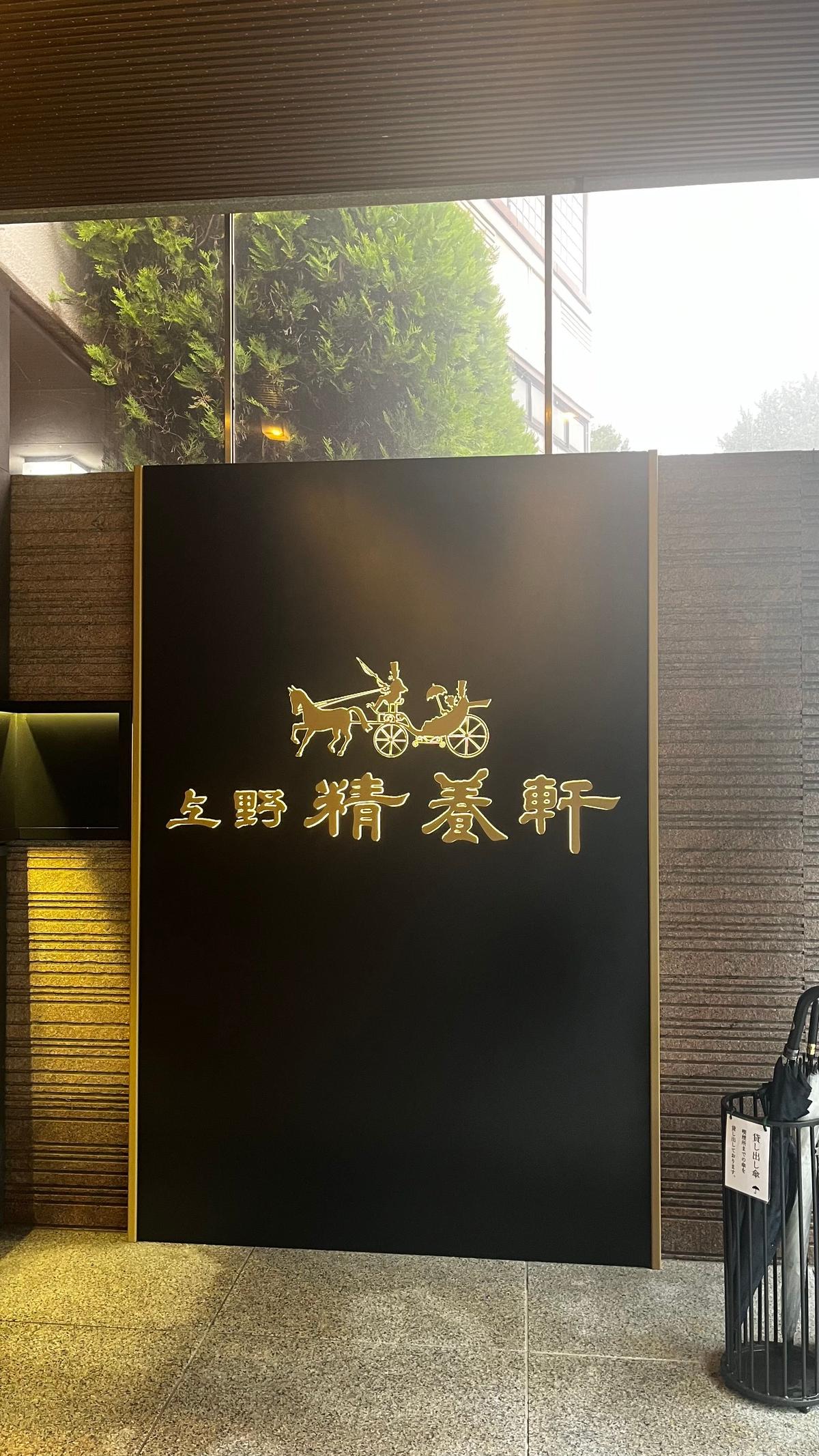 黒いパネルに、コーチのロゴマークと、漢字で「星野精養軒」の文字が金色で描かれている。背景には窓から見える緑の植栽。