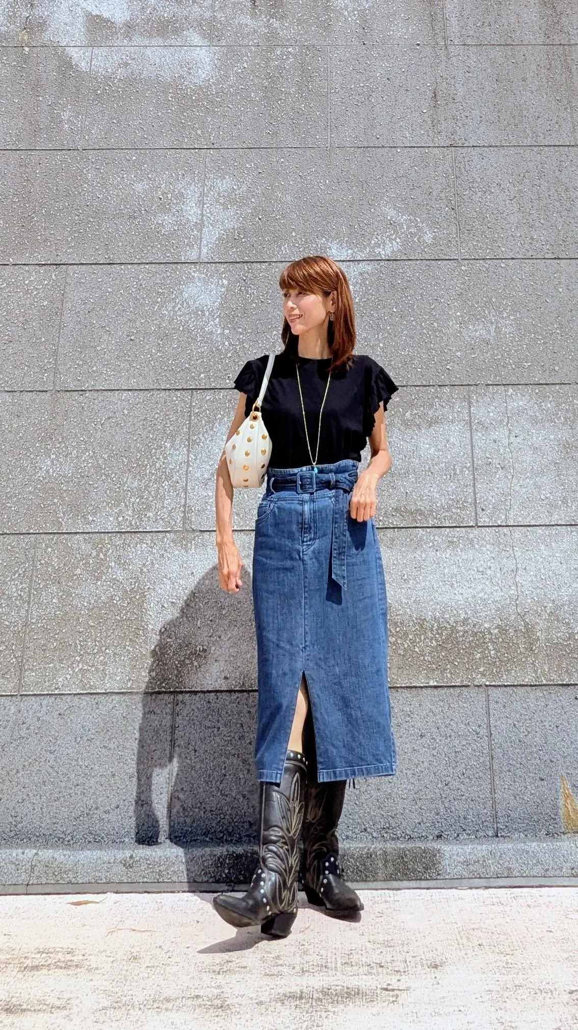 黒のトップスにデニムのミモレ丈スカート、黒い刺繍入りブーツを合わせた女性。白いショルダーバッグを斜め掛けし、腰にベルト。背景はグレーのコンクリート壁。