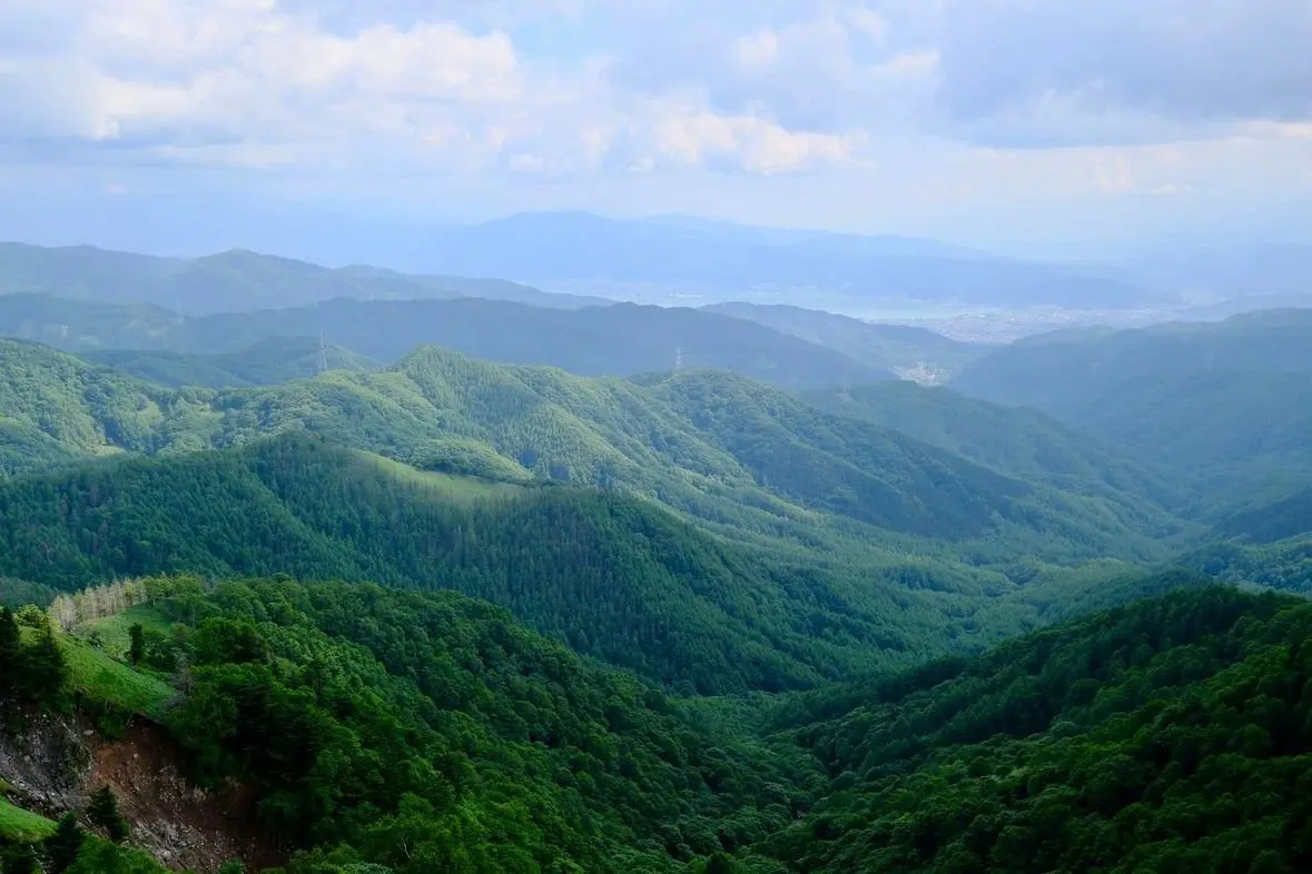 緑豊かな山々が連なり、遠景には霞んだ山並みが広がる風景。空は雲が浮かび、全体的に青と緑のコントラストが美しい。