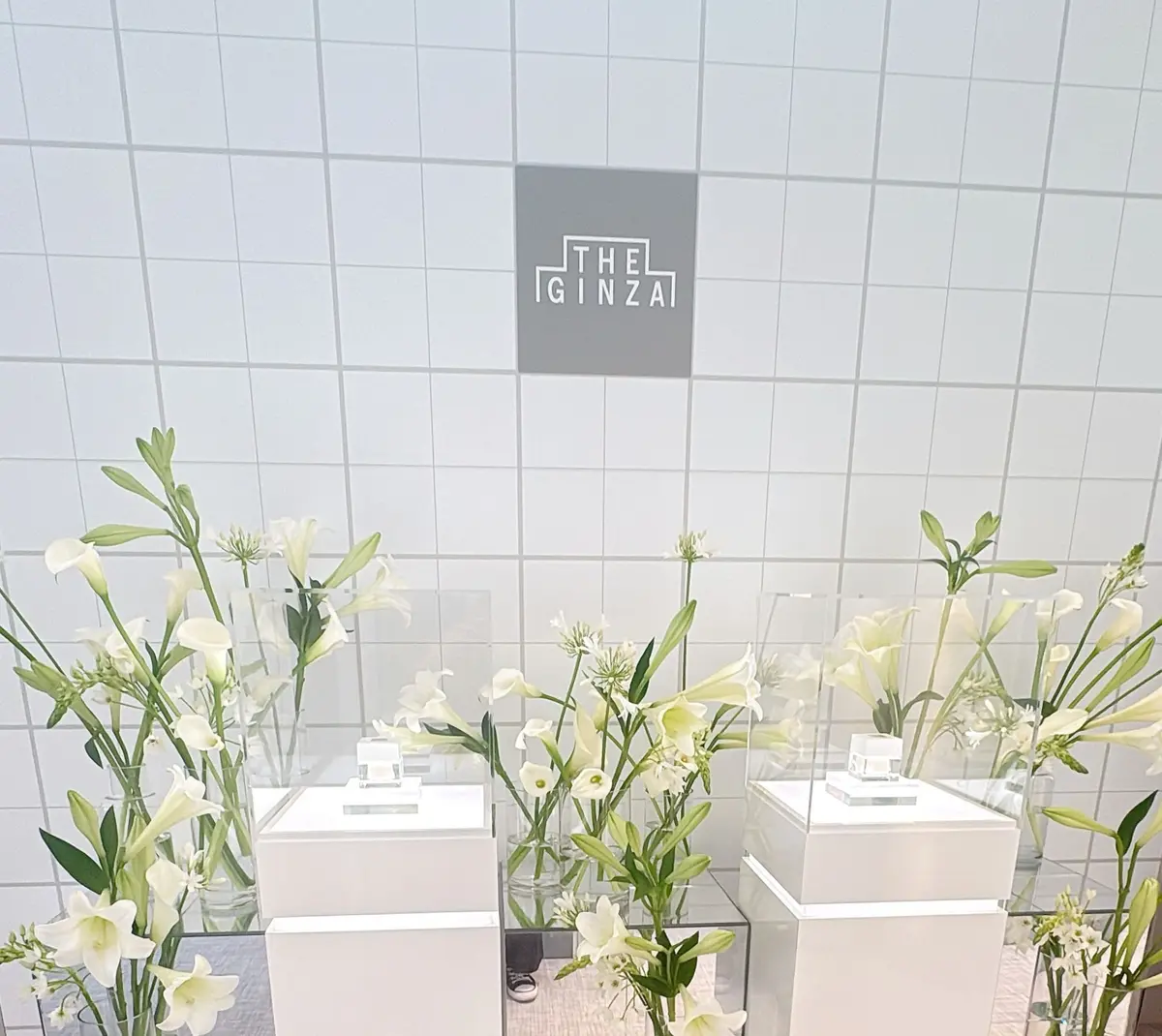 白いタイル壁の前に、白い花束と香水瓶が置かれている。中央には「THE GINZA」のロゴが入った黒い板がある。