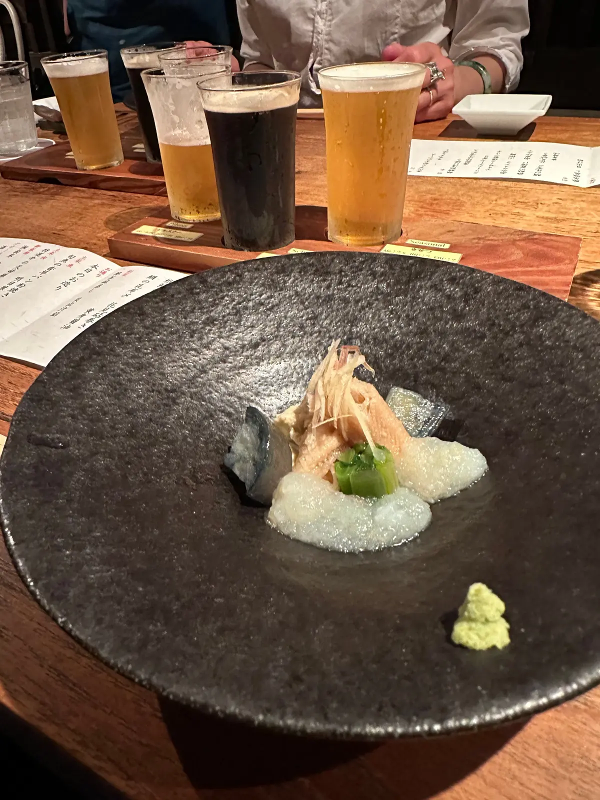 黒いプレートに、泡立つ白い液体、薄いピンクの魚の身、緑の野菜、青い海産物の盛り合わせ。