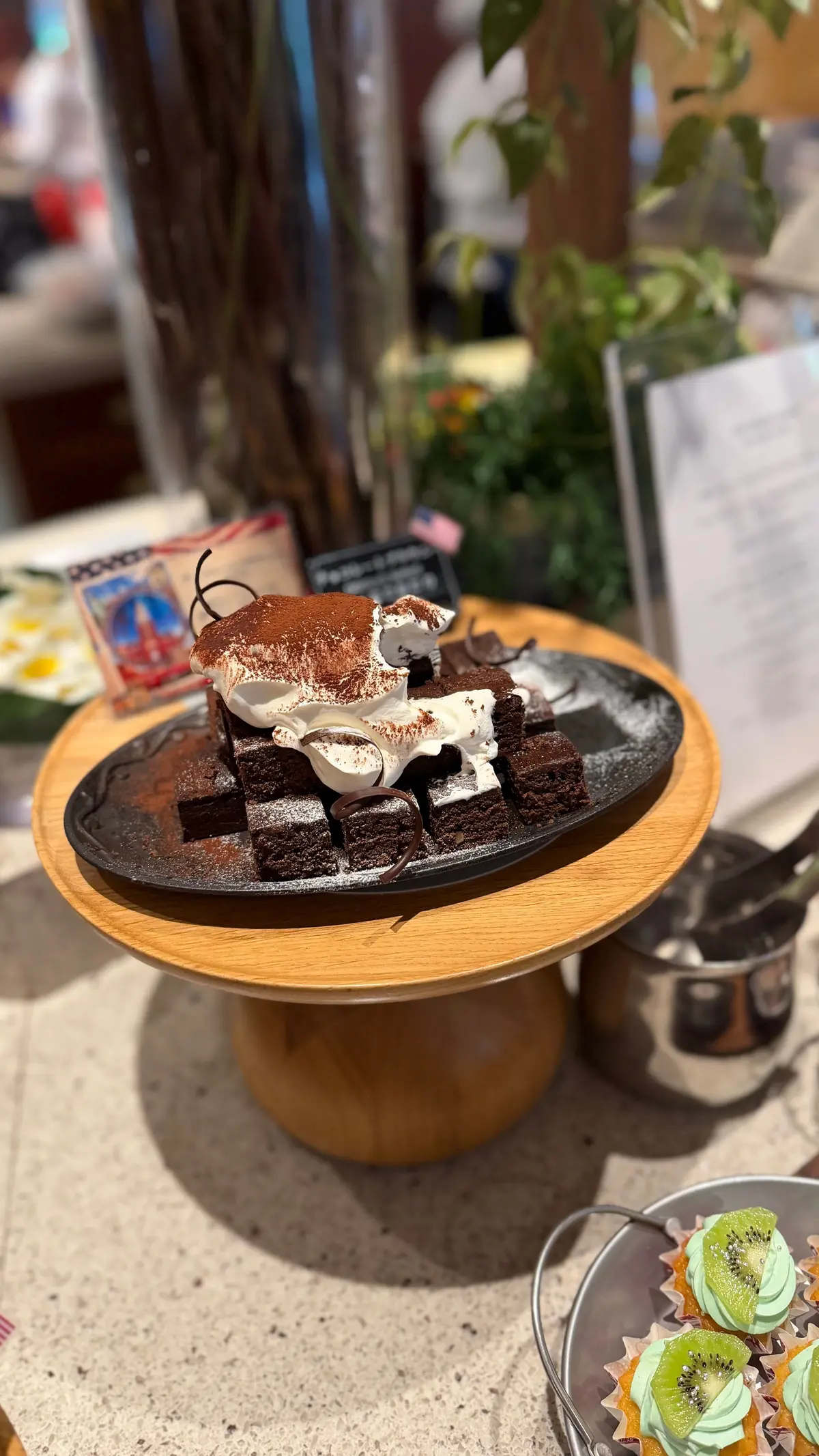 木製の台の上に置かれた、チョコレートケーキとブラウニーの盛り合わせ。ケーキは層状になっており、生クリームとココアパウダーがかかっている。ブラウニーは四角形でカットされ、ケーキの周りに配置されている。背景には植物やメニューなどがぼやけて見える。