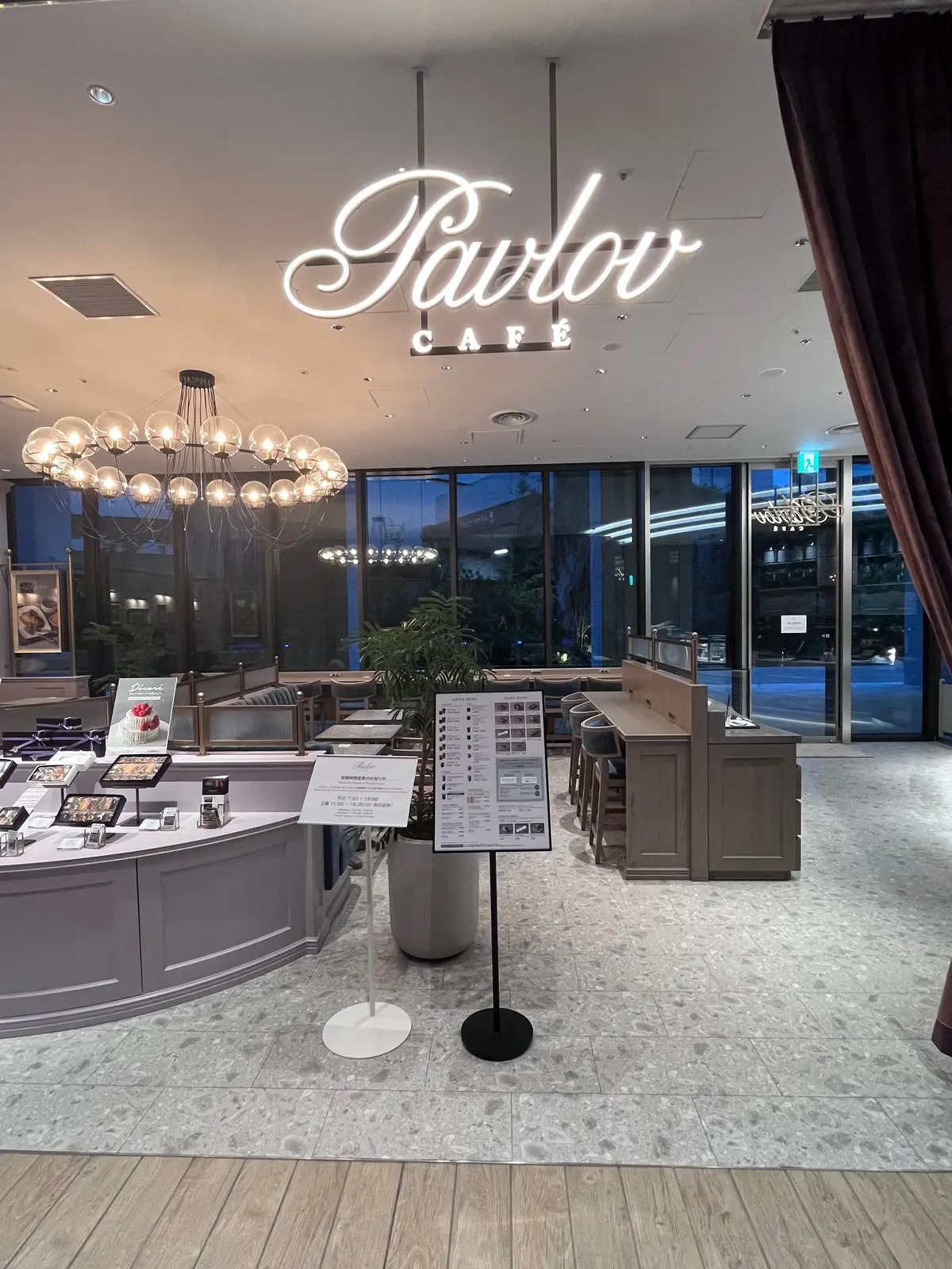 夜の Parlor カフェの店内。ネオンサインが輝き、大きな窓の外に街の灯りが広がる。中央にはメニュー台、左にはショーケースが置かれている。
