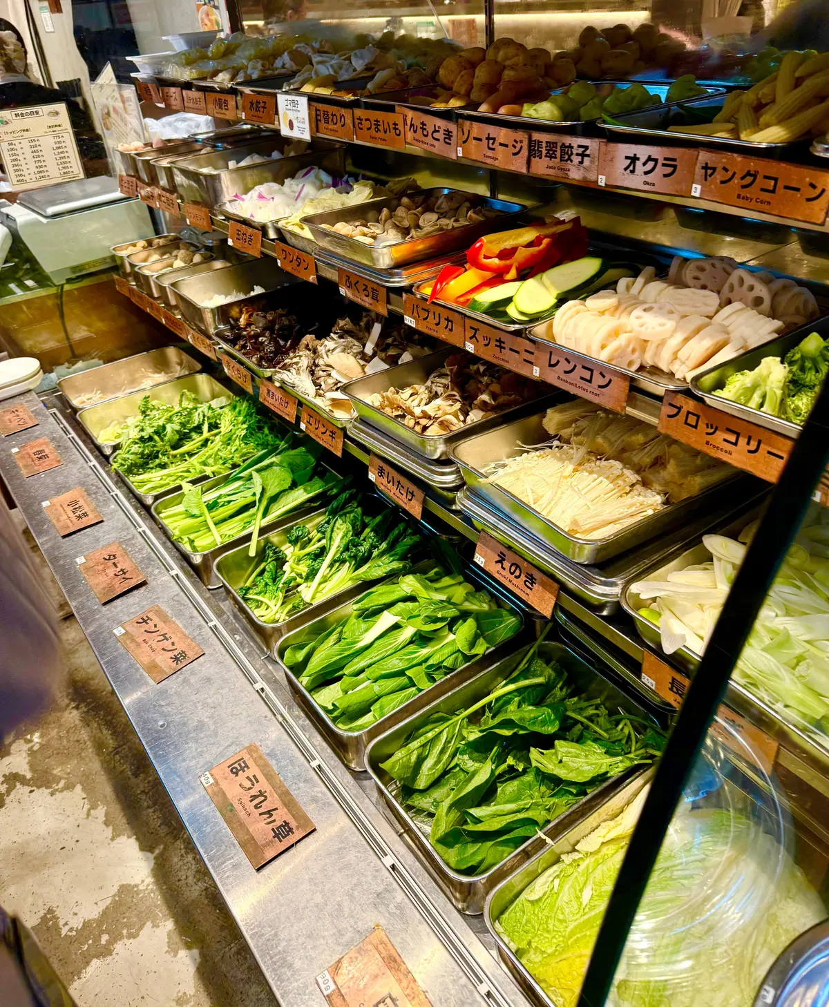 冷蔵ケースに並んだ、様々な種類の新鮮な野菜。緑色の葉物野菜、白い根菜、赤や黄色のパプリカなど、種類豊富に陳列されている。手書き風のラベルが各野菜に付けられている。