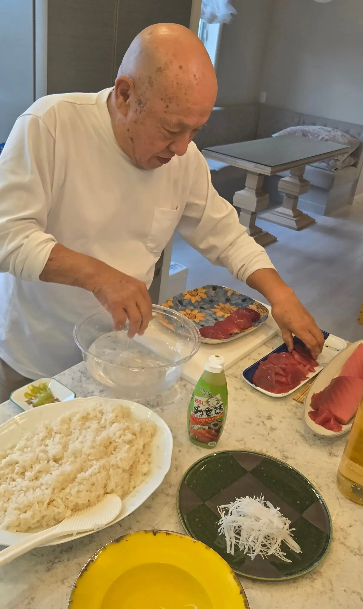 白髪の男性が、おにぎりを作る様子。手前にご飯、刺身、ワサビ、海苔などの材料が並び、男性は透明なボウルからご飯に何かを混ぜ込んでいる。