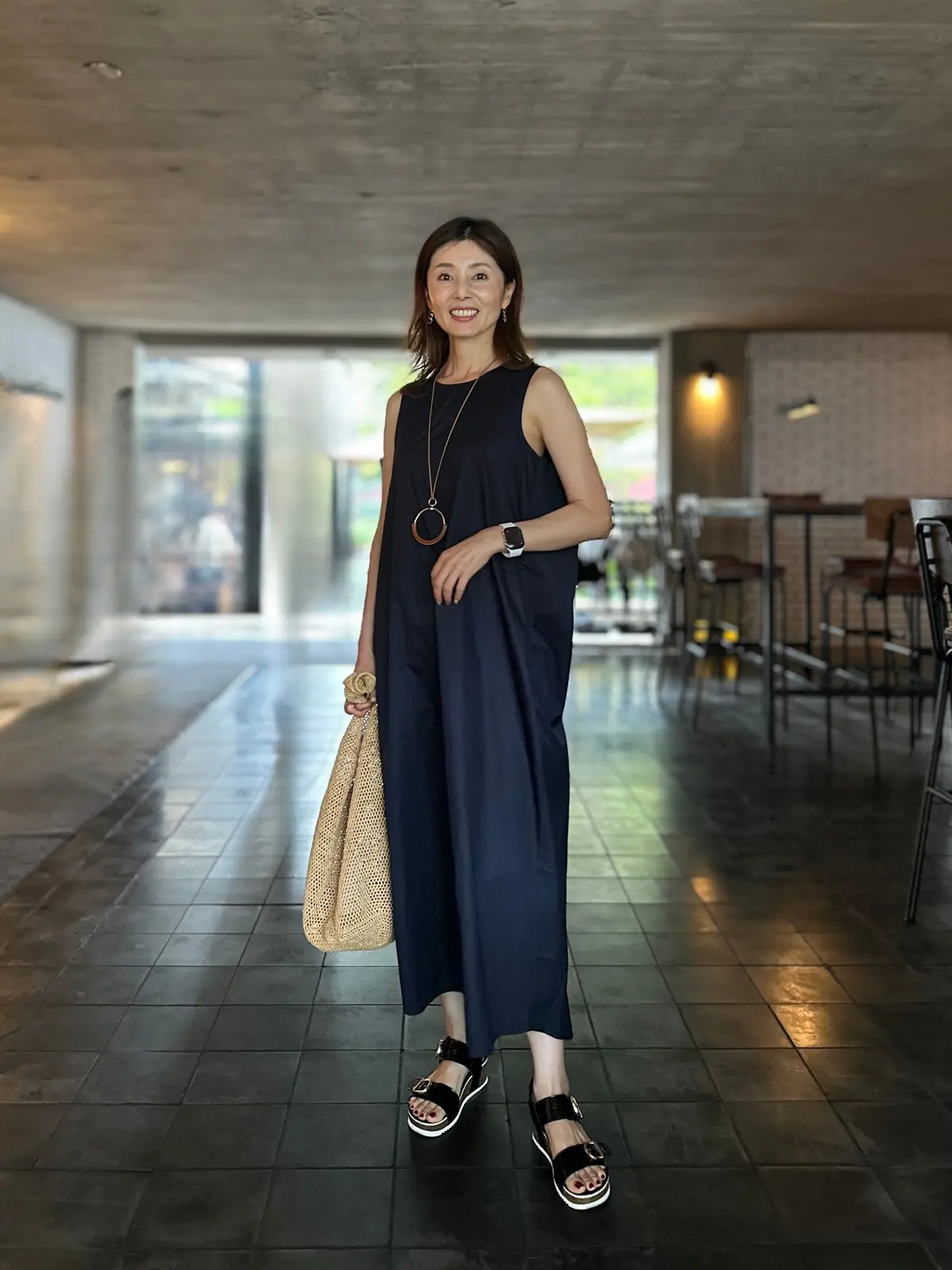 濃紺のワンピースを着た女性が、カフェのような空間で微笑んでいる。茶色の編みバッグを持ち、黒いサンダルを履いている。ネックレスとイヤリングを身につけ、落ち着いた雰囲気を醸し出している。