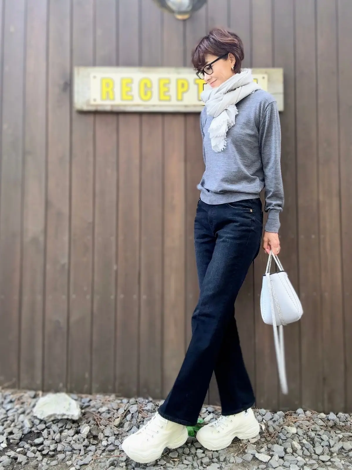 グレーのニットにデニム、白いスニーカーを合わせた女性。白いマフラーを巻いており、右手に白いハンドバッグを持っている。背景には「RECEPT」と書かれた看板がある木製の壁。