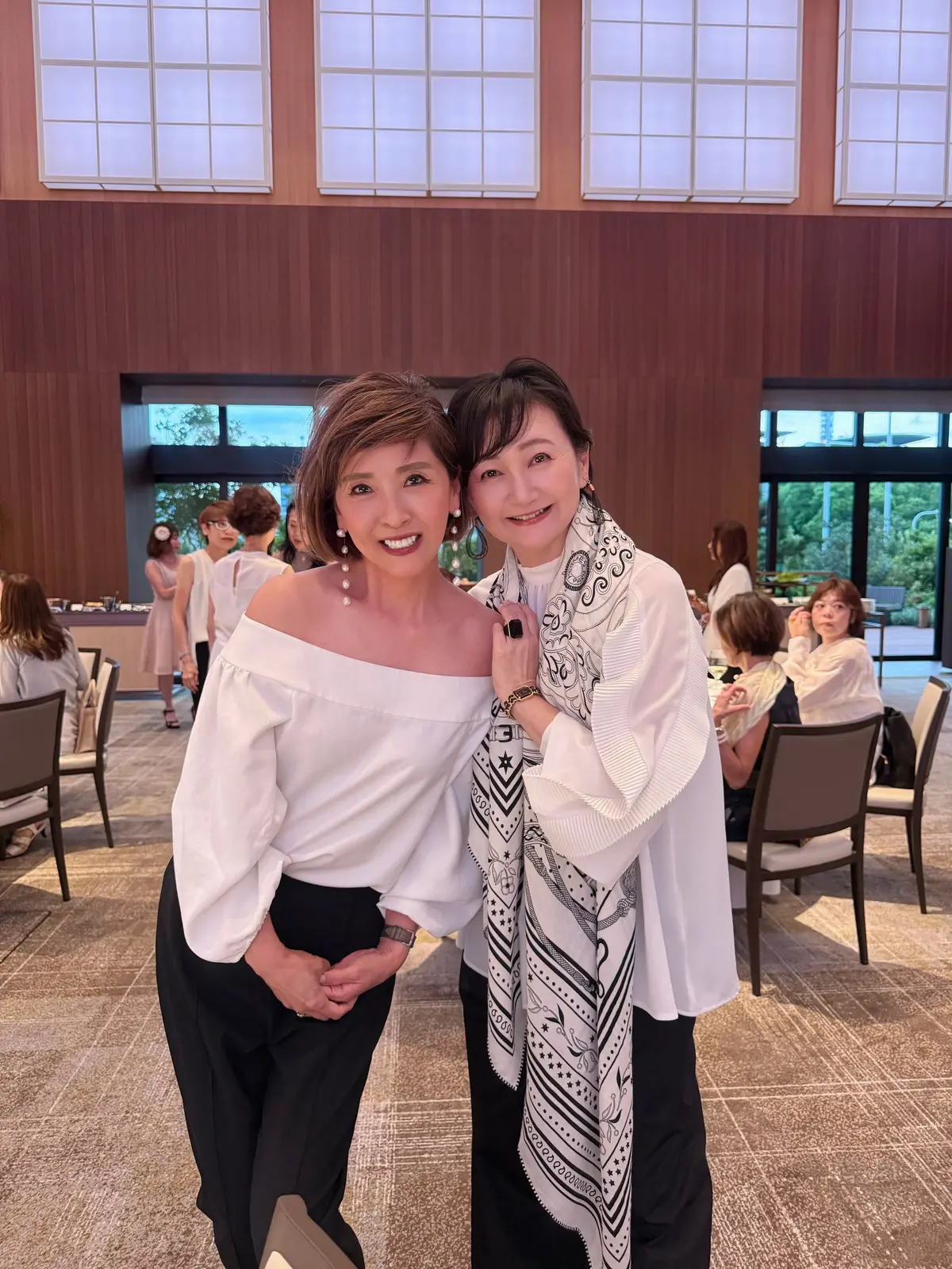 白シャツを着た女性と、柄物のベストを着た女性が笑顔で並んで立つ写真。背景には窓や木が見える明るい空間。