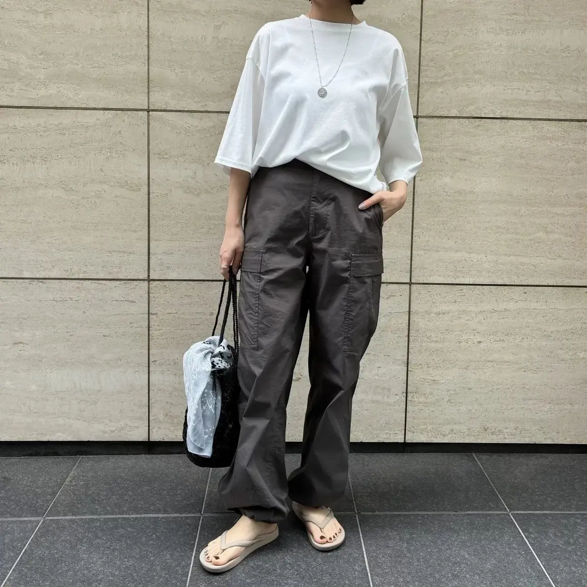 白Tシャツに茶色のカーゴパンツ、ベージュのサンダルを合わせた女性。黒いバッグを斜め掛けし、ネックレスを着用。背景はベージュのタイル壁。