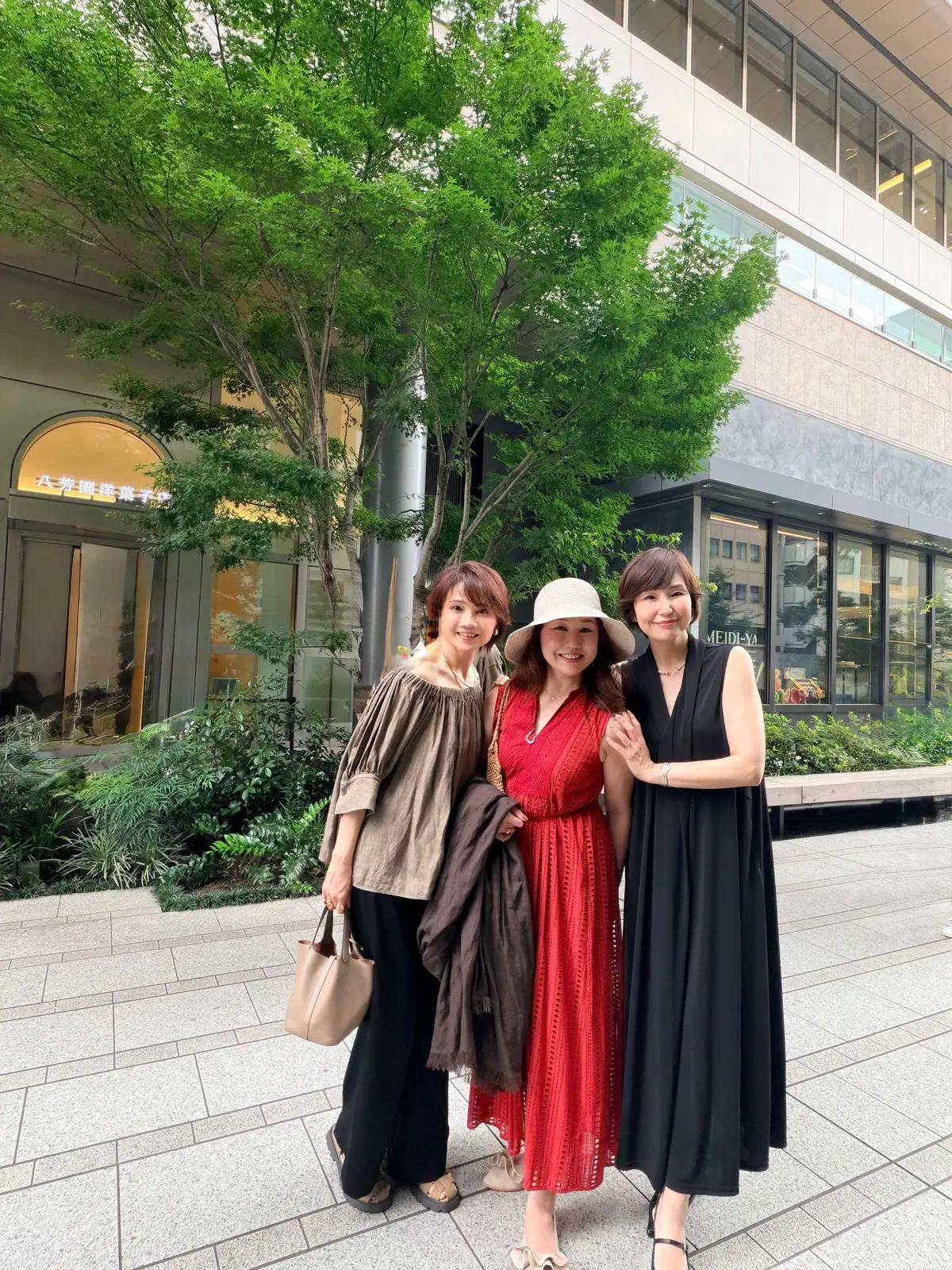 3人の女性が都市の歩道で並んで立っている。左から、緑色のトップスと黒いパンツ、茶色の肩掛け、右から、赤い花柄のワンピースに帽子、黒いワンピース。背景には建物と木々が見える。