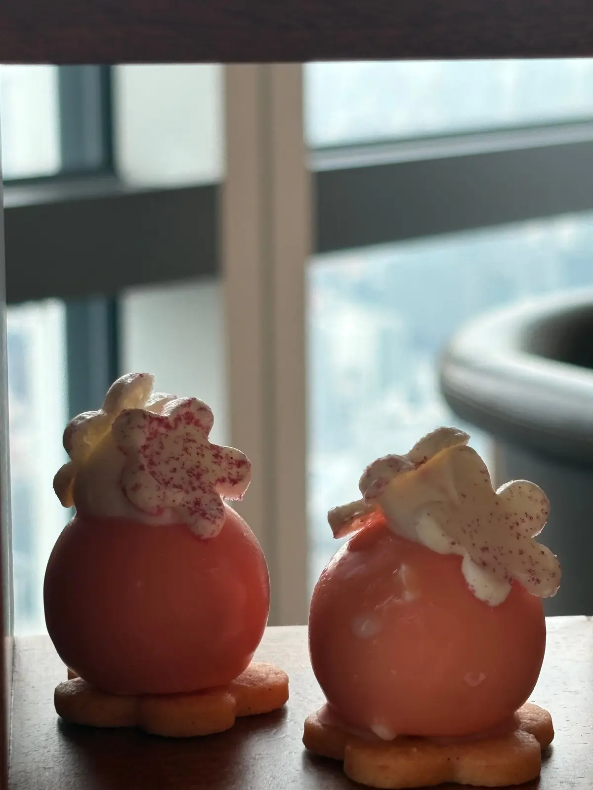 ピンク色の球体スイーツ2つが木製の台に並んでいる。それぞれにクリームと赤い粉末をかけた飾りがあり、下には薄いクッキーが敷かれている。背景には窓からの光が差し込んでいる。
