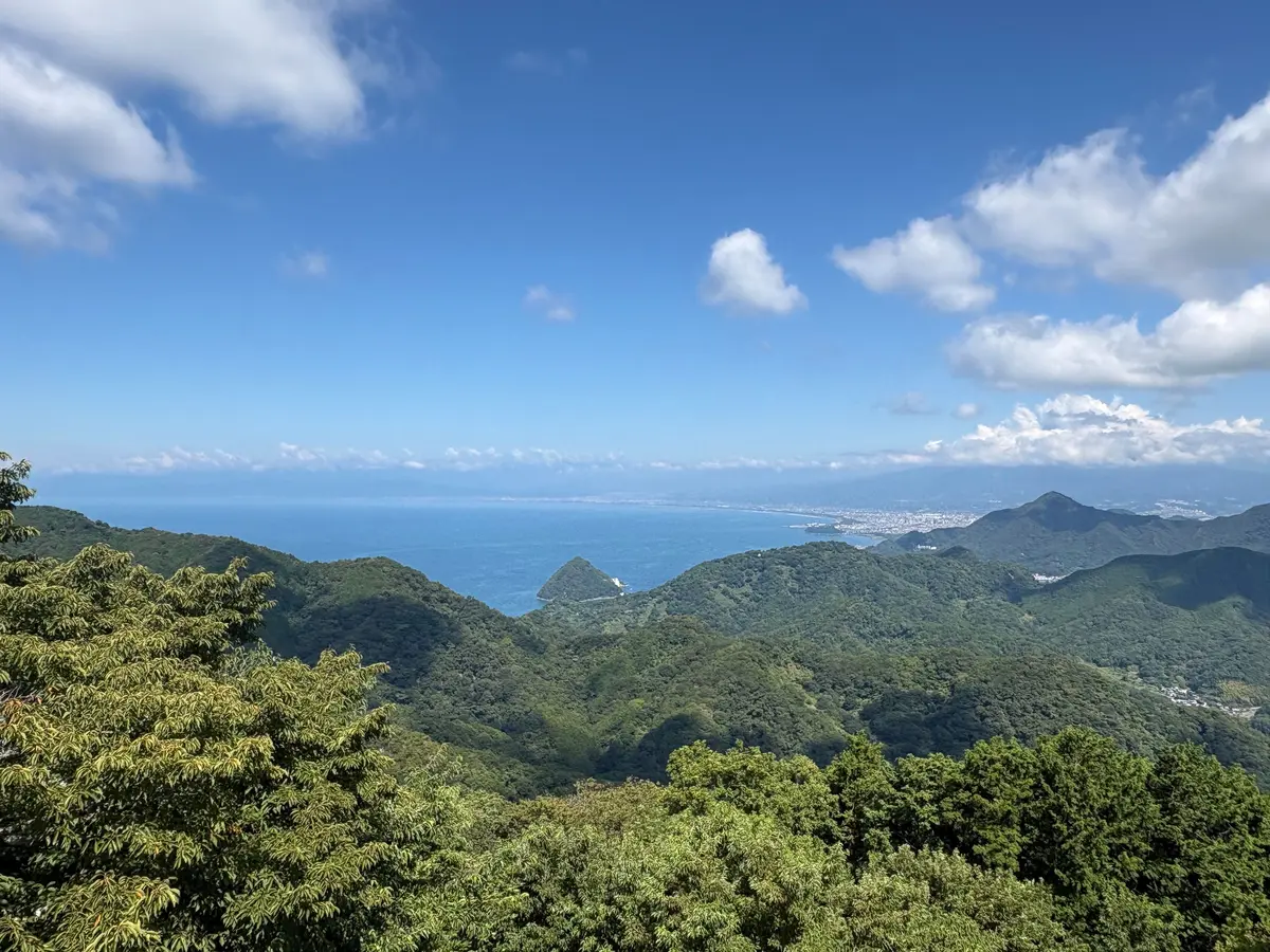 緑豊かな山々が連なり、遠くに青い海と海岸線が見える風景。晴天の空には白い雲が浮かんでいる。