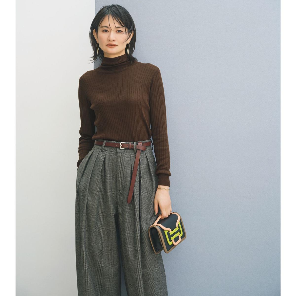 茶色のリブニットのタートルネックセーターに、グレーのチェック柄パンツを合わせた女性。茶色のベルトがウエストに巻かれ、ゴールドのピアスとブレスレットを着用。右手に赤いバッグを持つ。背景は白とグレーの壁。