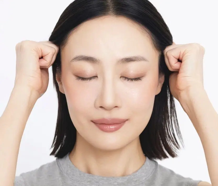 側頭筋や前頭筋をほぐし、 重しとなる老廃物を流す　女性が両手で耳を押さえて、目を閉じています。グレーのトップスを着ています。