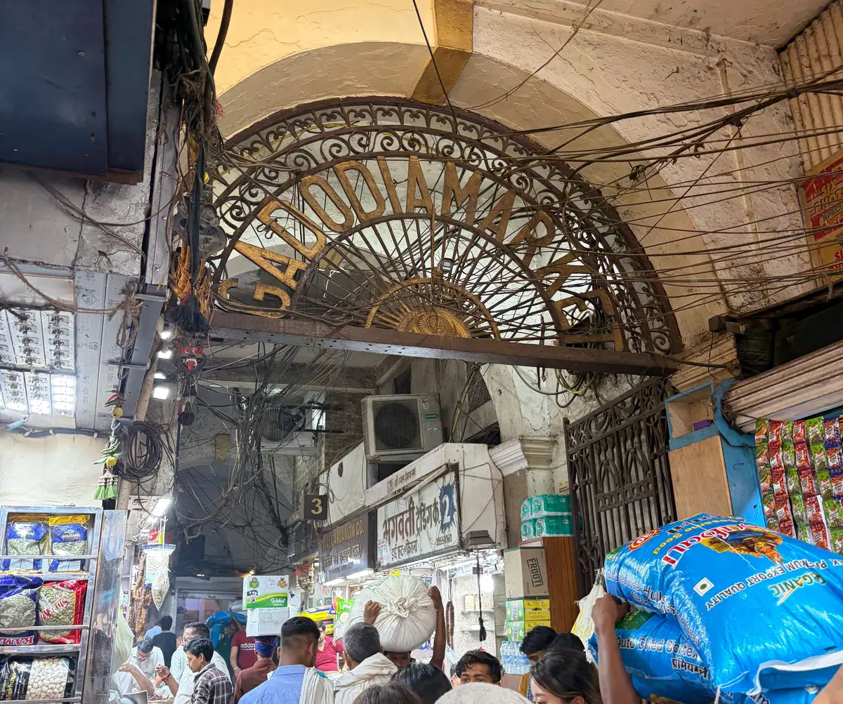 インドの市場の入り口。アーチ型の装飾が施されたゲートがあり、その向こうに多くの人々が歩いている。周囲には店が並び、電線が複雑に張り巡らされている。
