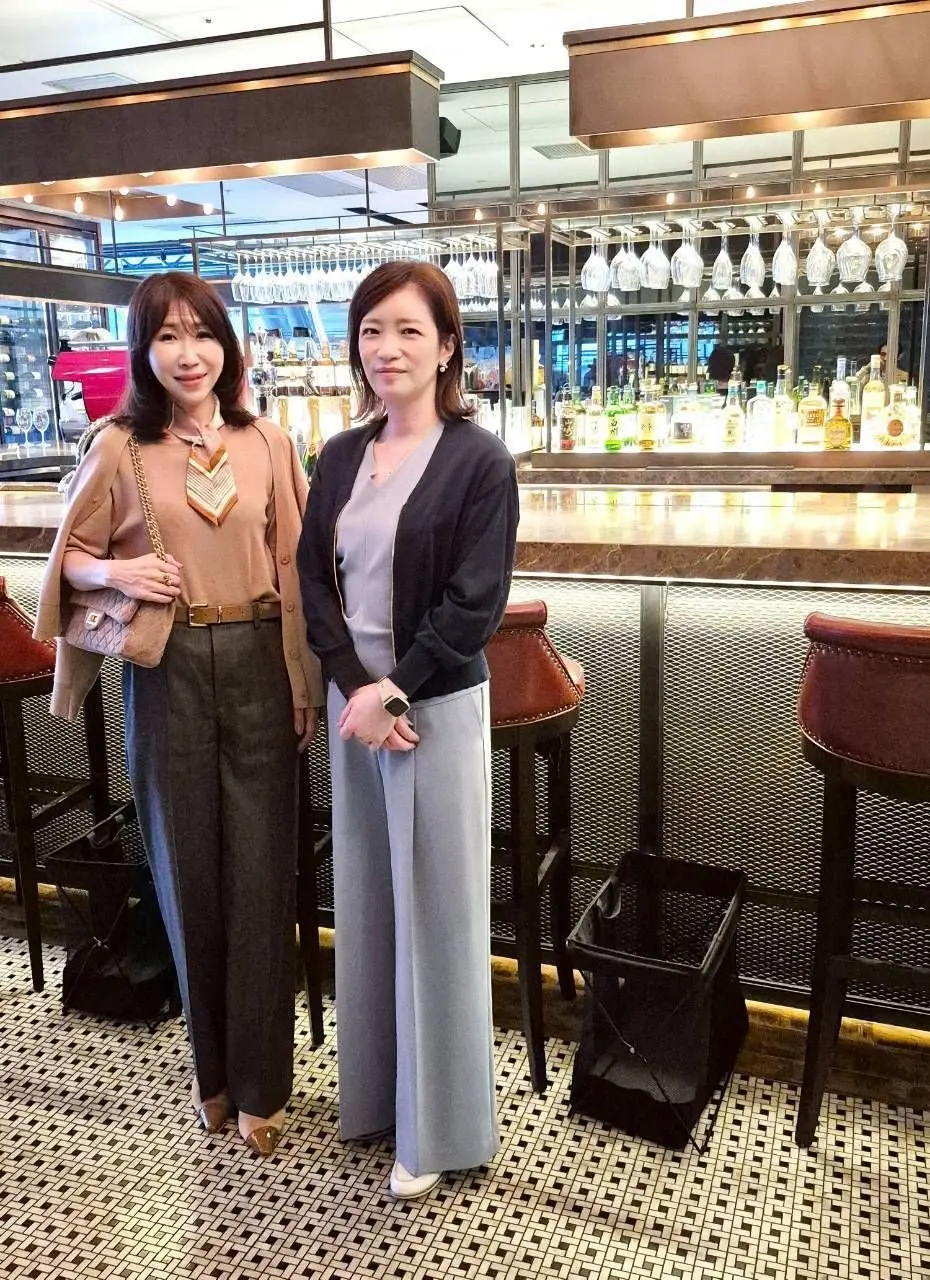 バーカウンターに立つ2人の女性。左はベージュのジャケットに黄色の襟付きシャツ、右はグレーのカーディガンにワンピースを着ている。背景には棚にグラスやボトルが並び、モダンな内装が見える。