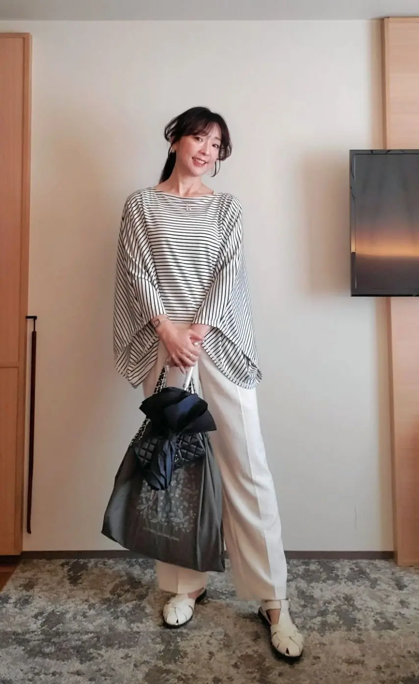 白いカーペットの上で、女性が笑顔で立っている。黒と白のボーダーのトップスに、白いゆったりとしたパンツを着用。グレーの大きなトートバッグを持ち、白い編み込みサンダルを履いている。背景には木製の家具とテレビがある。