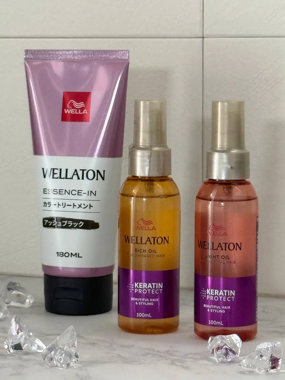 左から、ウェルアトン（Wellaton）のエッセンスインカラートリートメント、リッチオイル、ライトオイルの3つのヘアケア製品が並んでいる。ピンク、オレンジ、茶色のボトルで、それぞれ180ml、100mlの容量。背景にはクリスタルのような装飾がある。