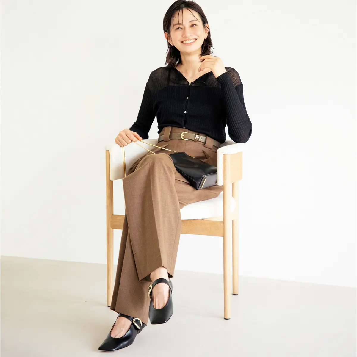 ベージュのワイドパンツに黒のトップス、黒いフラットシューズを着用した女性が椅子に座っている全身写真。金色のベルトを締め、笑顔でこちらを見ている。背景は白。