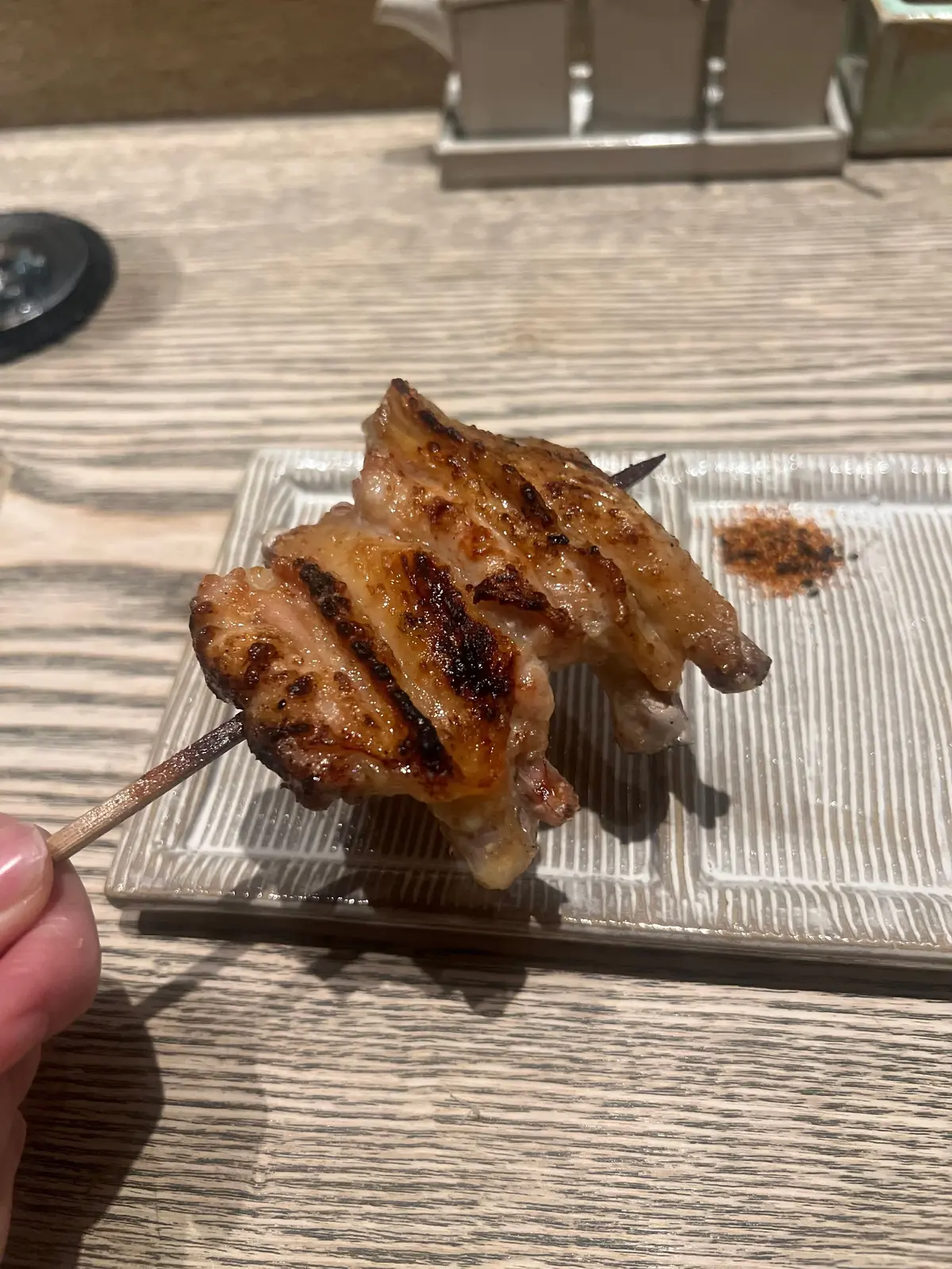 焼き鳥の串焼きが白い長方形の皿に載せられ、木製の串で支えられている。表面は焦げ目があり、木製のテーブルに置かれている。