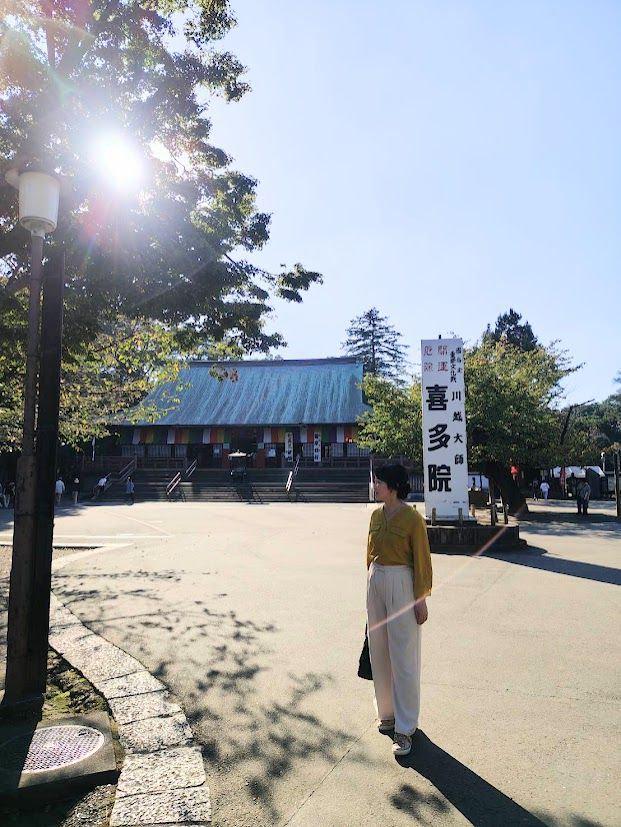 広々とした石畳の広場に立つ女性。背景には、朱色の屋根の伝統的な建物と、そこに掲げられた「喜多院」と書かれた看板が見える。晴天で太陽が強く、光が広場に降り注いでいる。