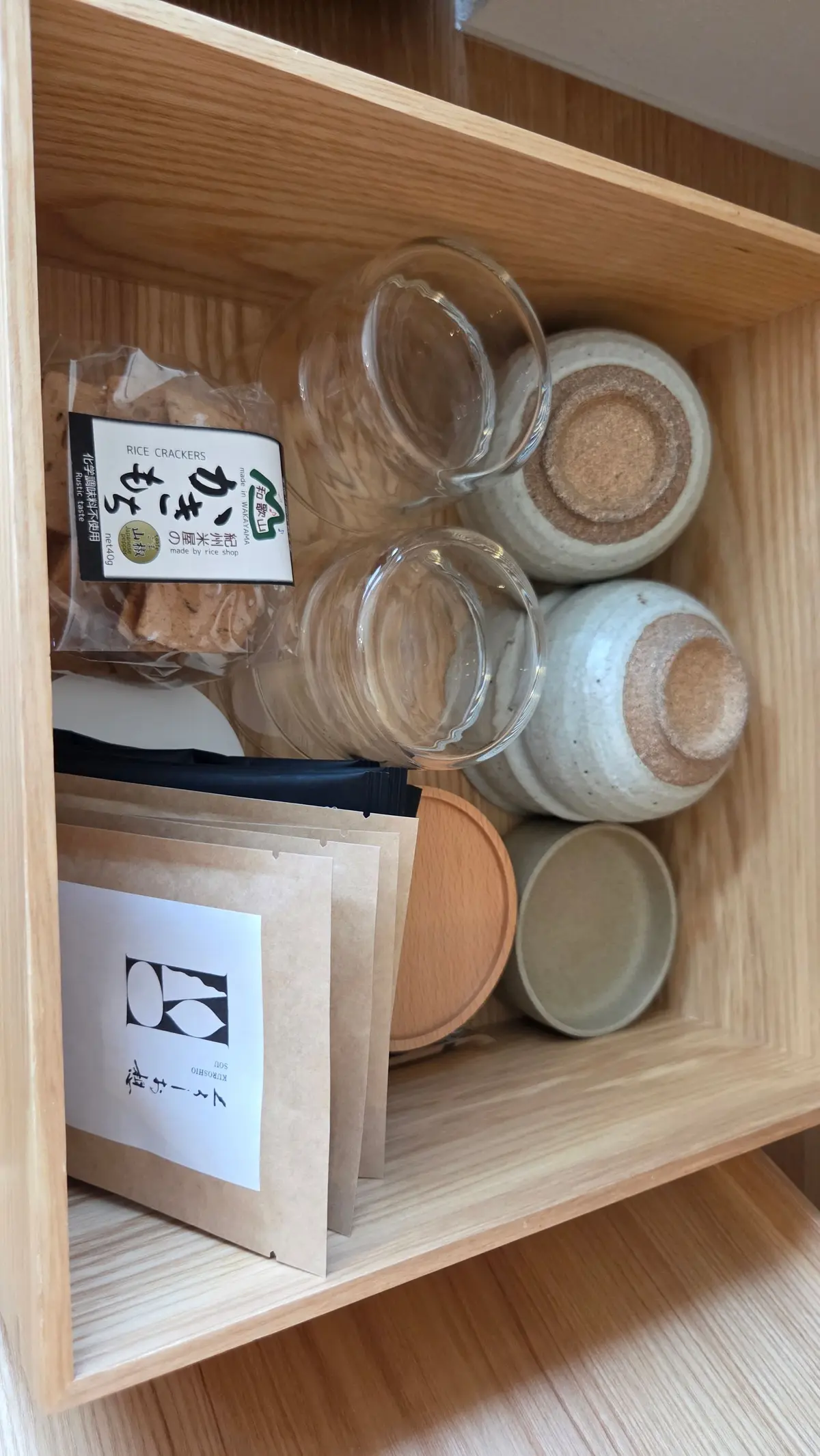 木製の棚に、米菓の瓶や箱が並んでいる。瓶には茶色い米クラッカーが入っており、ラベルが貼られている。箱は白地に黒の文字で「ふくや」と記載されている。