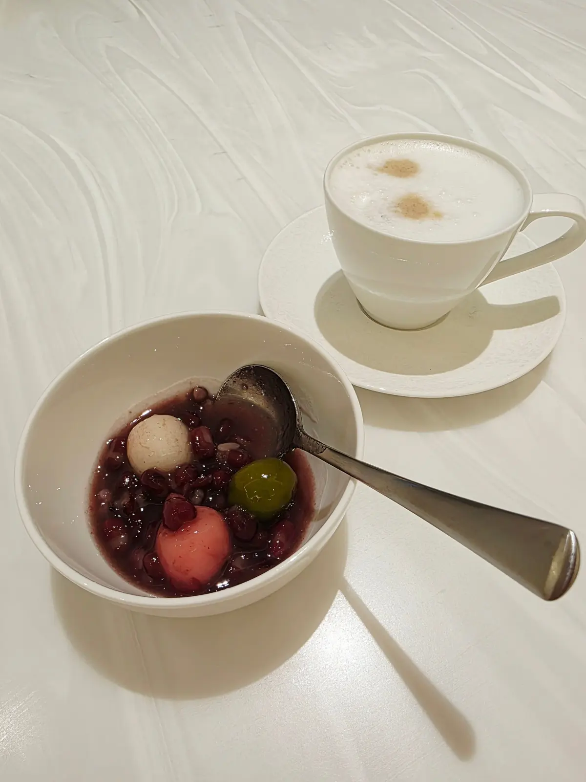 白いテーブルに置かれた、赤色の豆と白玉、そして緑色の柑橘類が入った器と、ミルクフォームが乗った白いカップ。スプーンが器に添えられている。