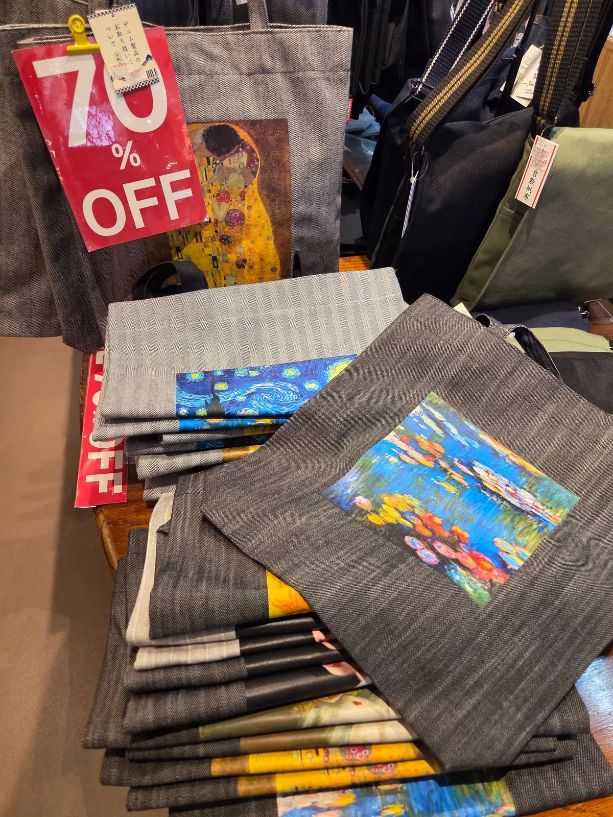 美術館の絵画をプリントしたグレーのトートバッグが並ぶ様子。70%OFFの赤いポップが立てかけられ、背景には黒いリュックサックも見えます。