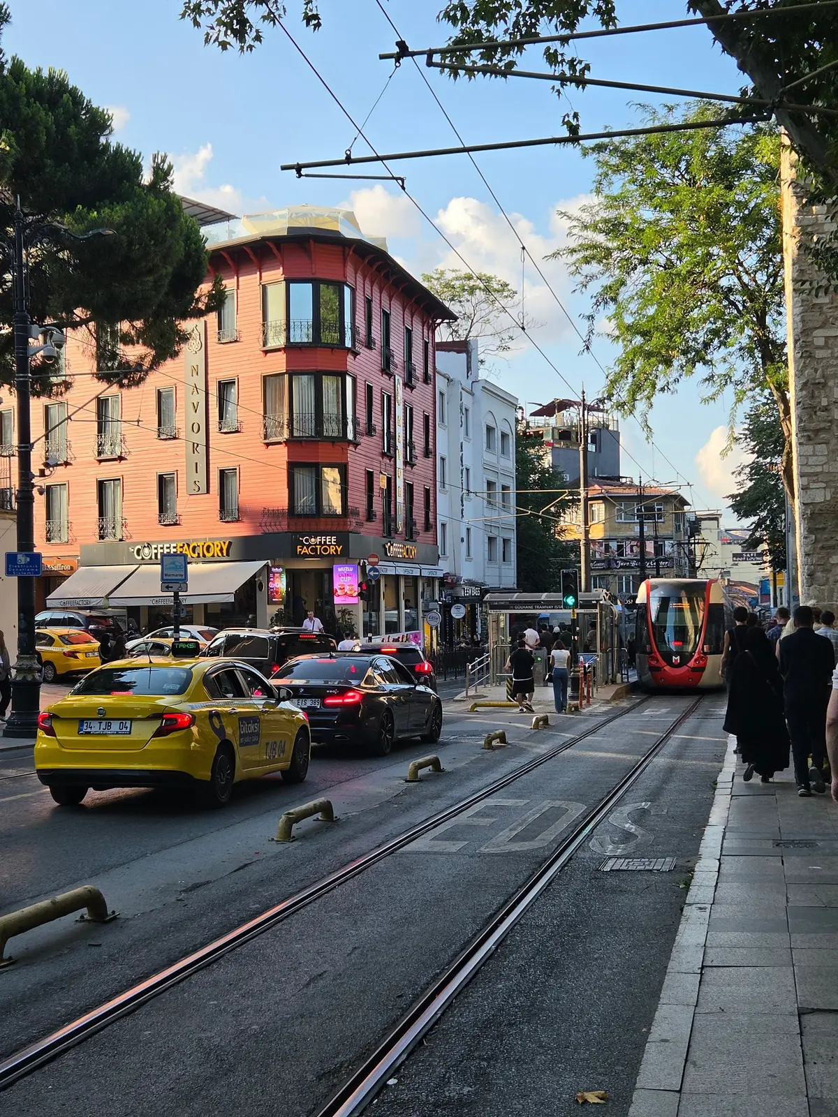 イスタンブールの街並み。オレンジ色の建物が目立つ。トラム線が走り、トラムが停車している。道路には黄色いタクシーを含む車が走る。歩行者もおり、賑やかな様子。