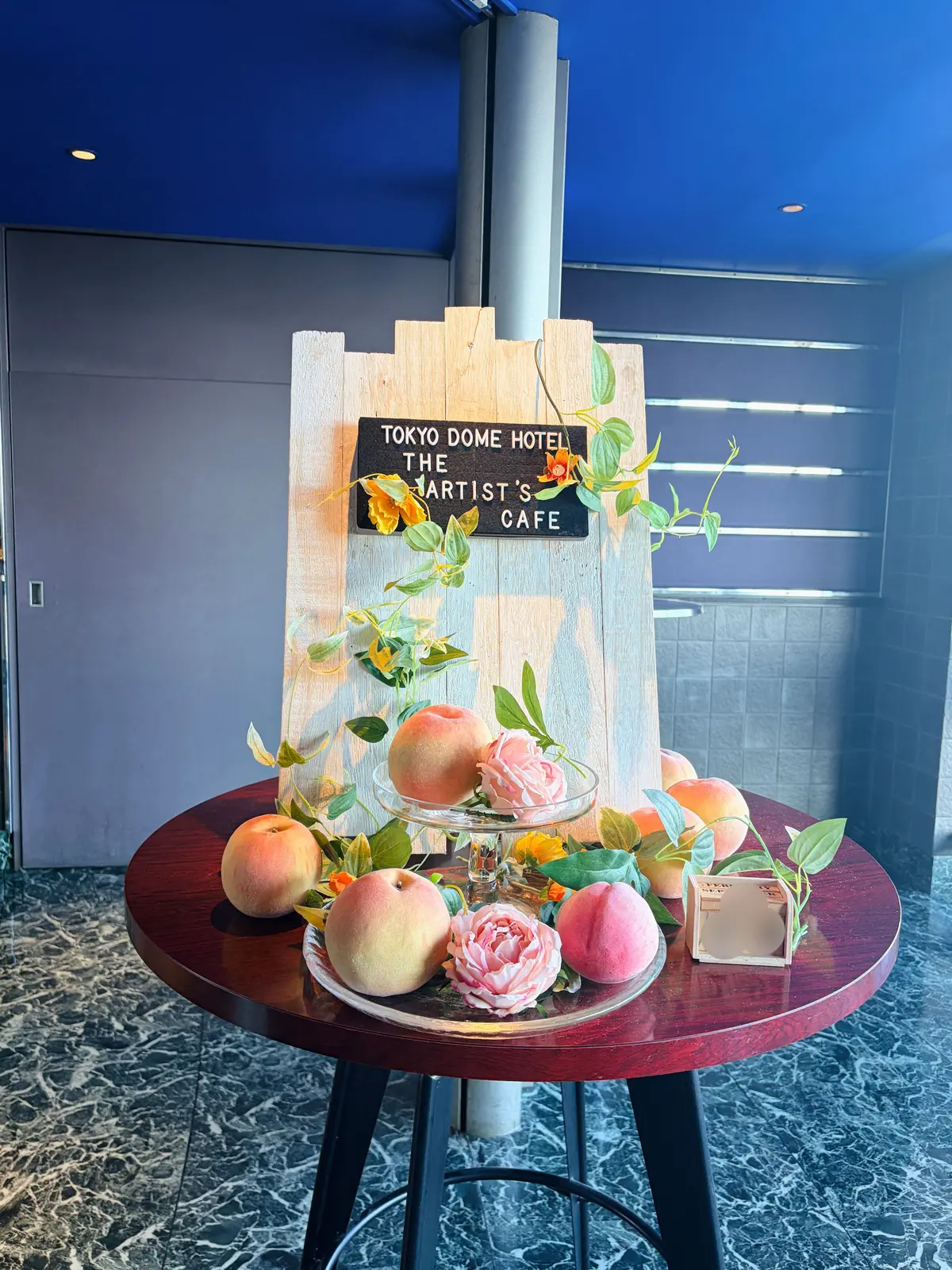 丸いテーブルに、桃や花、葉を飾った盛り合わせ。背景には「TOKYO DOME HOTEL THE ARTIST’S CAFE」と書かれた木製の看板。