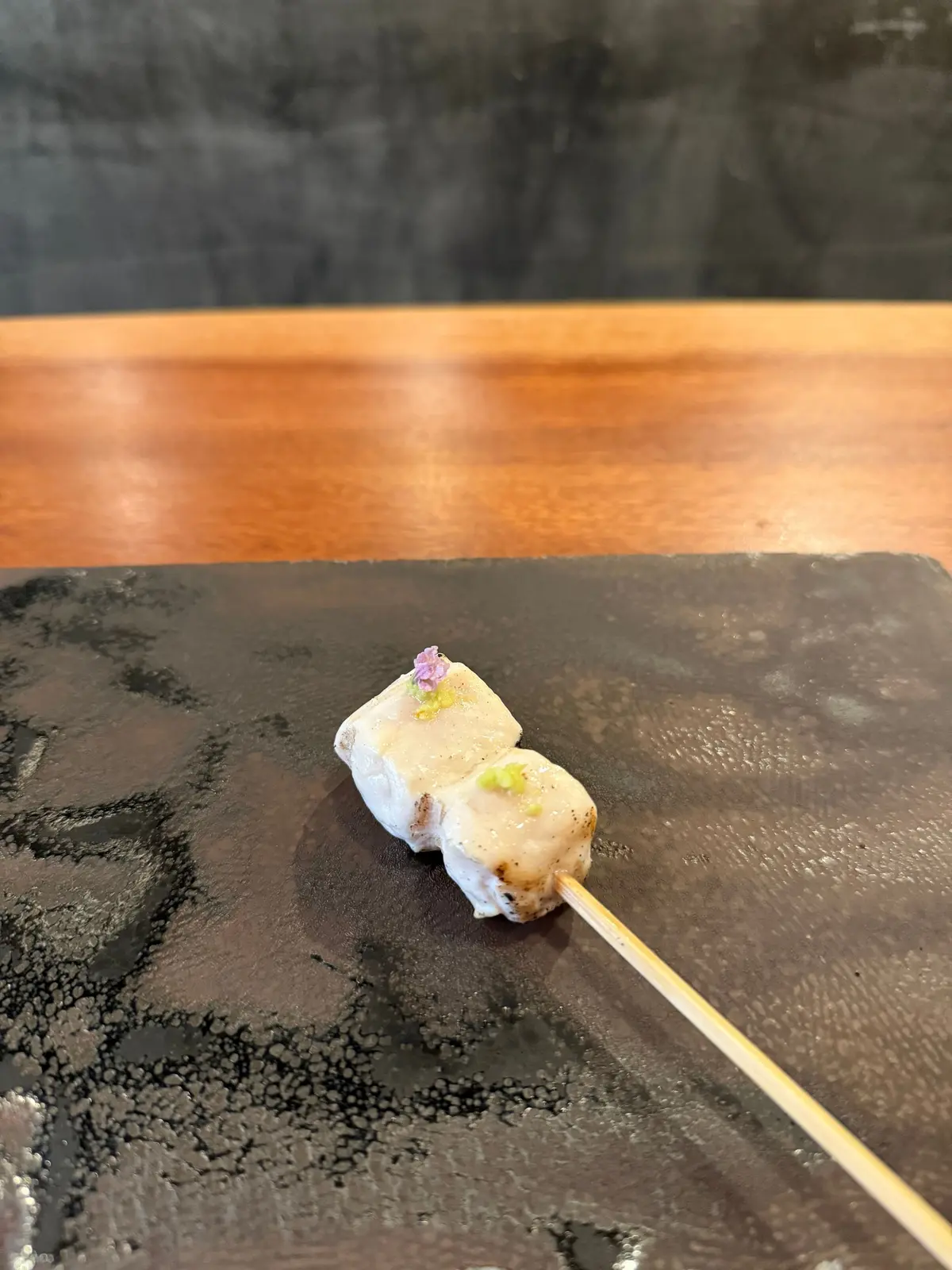 串に刺さった白身魚の焼き物。表面は軽く焦げ付き、ピスタチオと紫色の小さな花が添えられている。黒いお皿に置かれ、木の串が右下に向かって伸びている。
